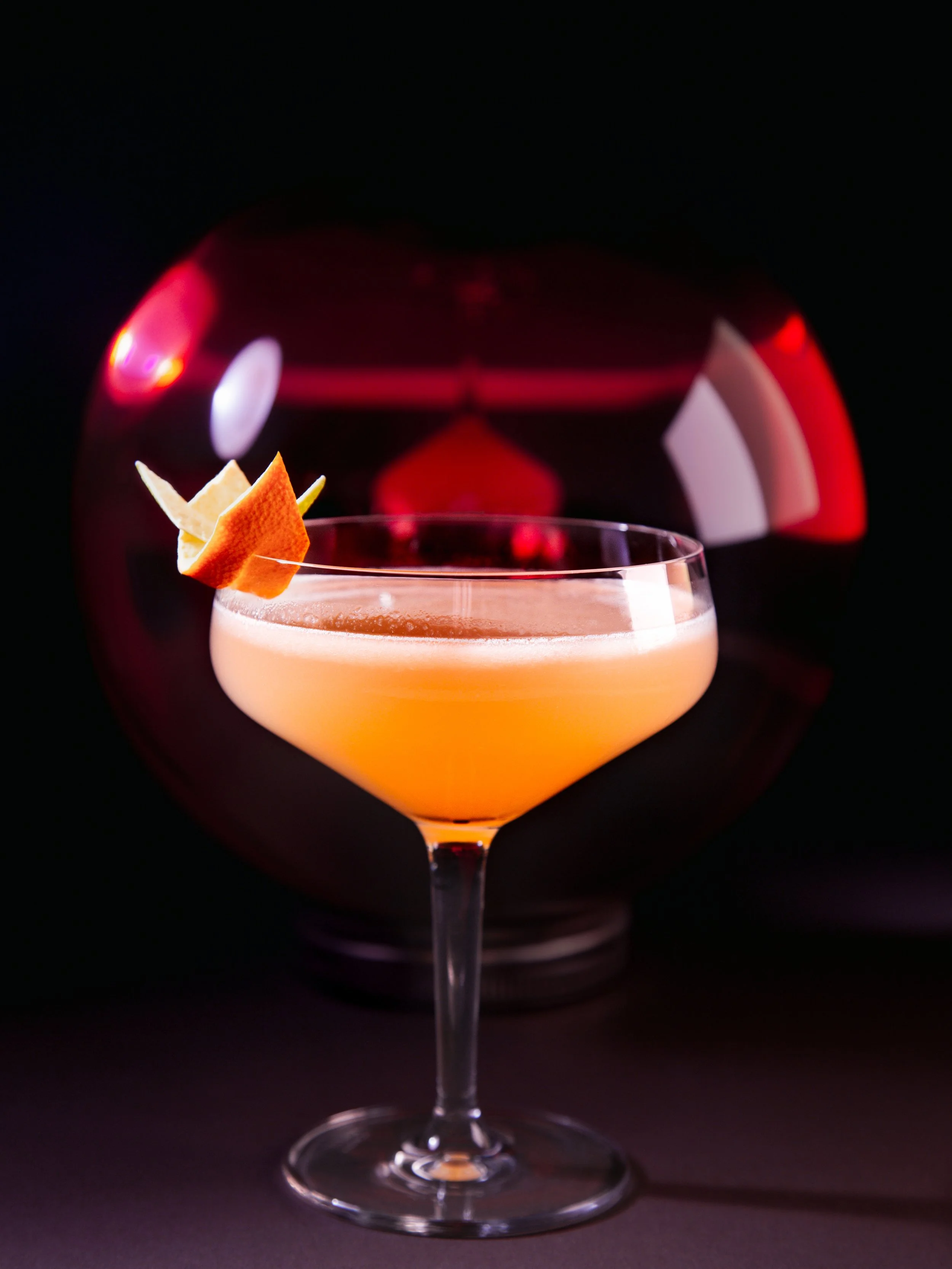 A cocktail glass with a creamy drink garnished with fruit and a peel, with a dark background and a red, blurred sphere in the background.