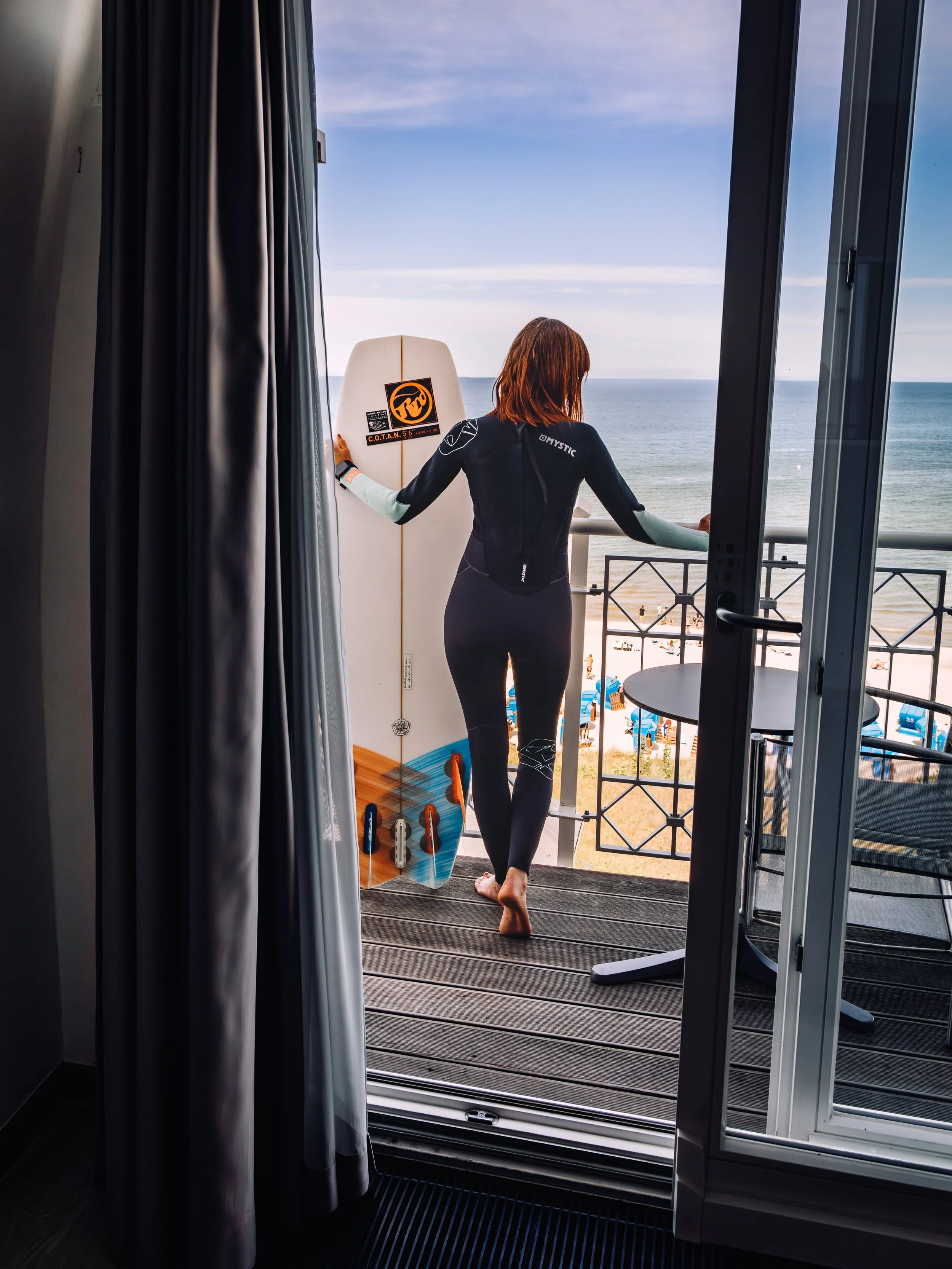 A woman in a wetsuit standing on a balcony, holding a surfboard with a sunset beach view in the background.