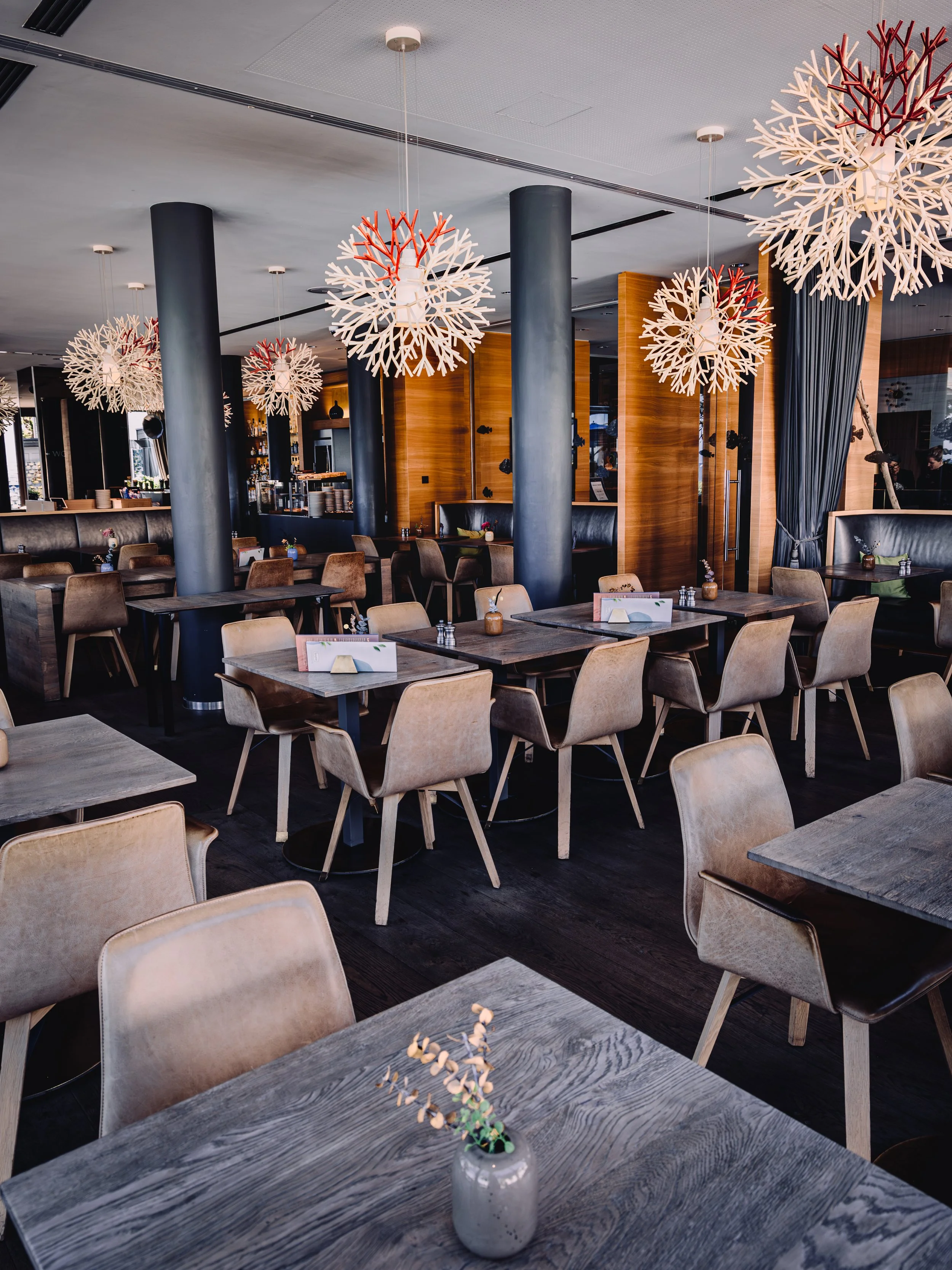 Interior of a modern restaurant with wooden tables, beige chairs, large black columns, and decorative white light fixtures resembling snowflakes hanging from the ceiling. The tables have small floral centerpieces in vases.
