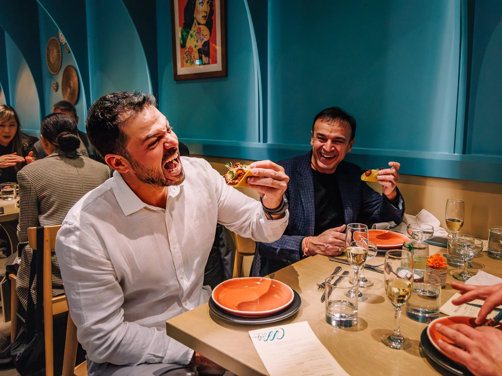 Two men laughing and enjoying food at a restaurant table, with dishes and glasses of water and wine, in a vibrant, decorated space with teal walls and artwork.