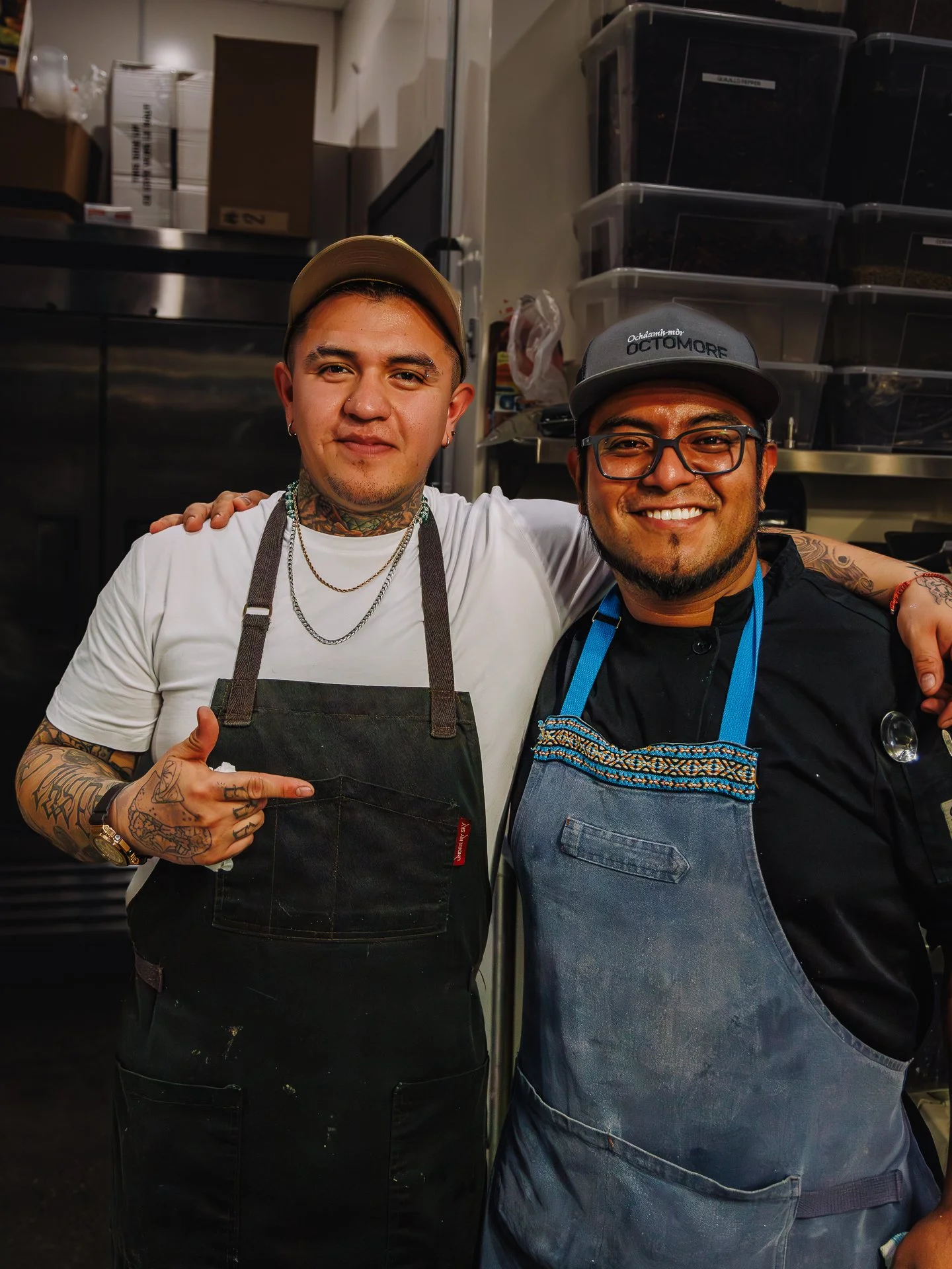 Two men standing inside a kitchen, smiling and posing for the camera. One man has tattoos and is wearing a light-colored cap, a white shirt, and a black apron. The other man has glasses and a dark cap, a black shirt, and a blue apron. The background 