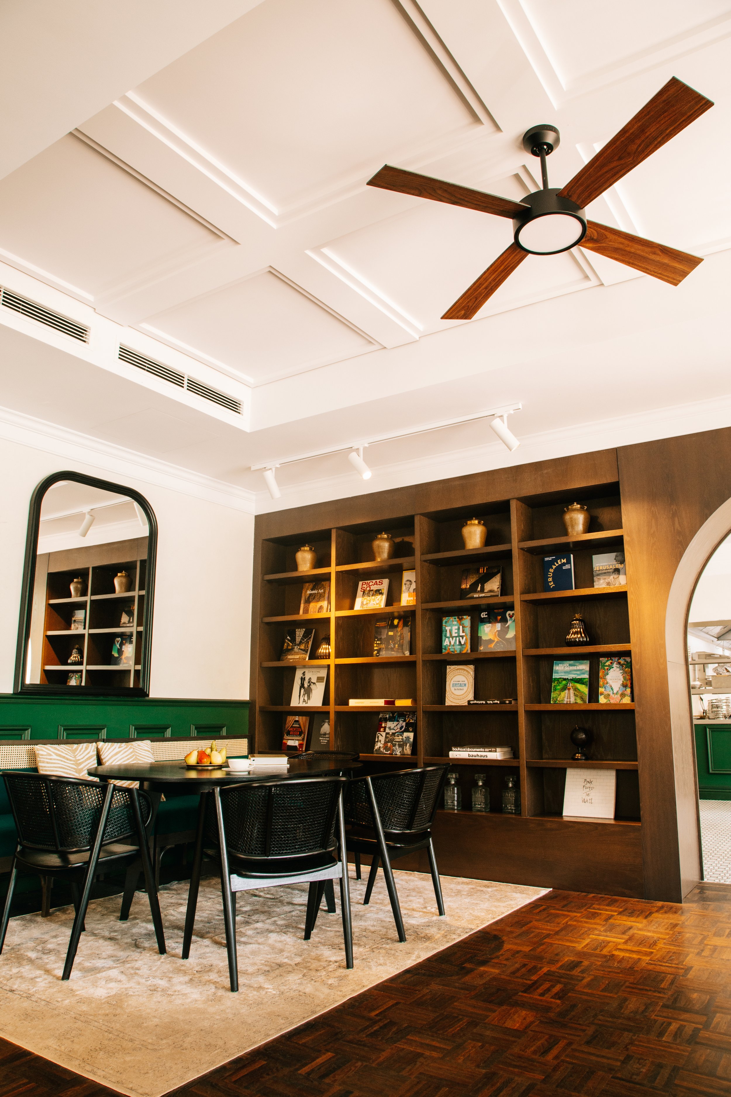 A modern dining room with a wooden bookshelf filled with books and decorative items. A round black dining table with black chairs is in front of a green accent wall with a mirror. The ceiling has a contemporary black and wood ceiling fan, and there a