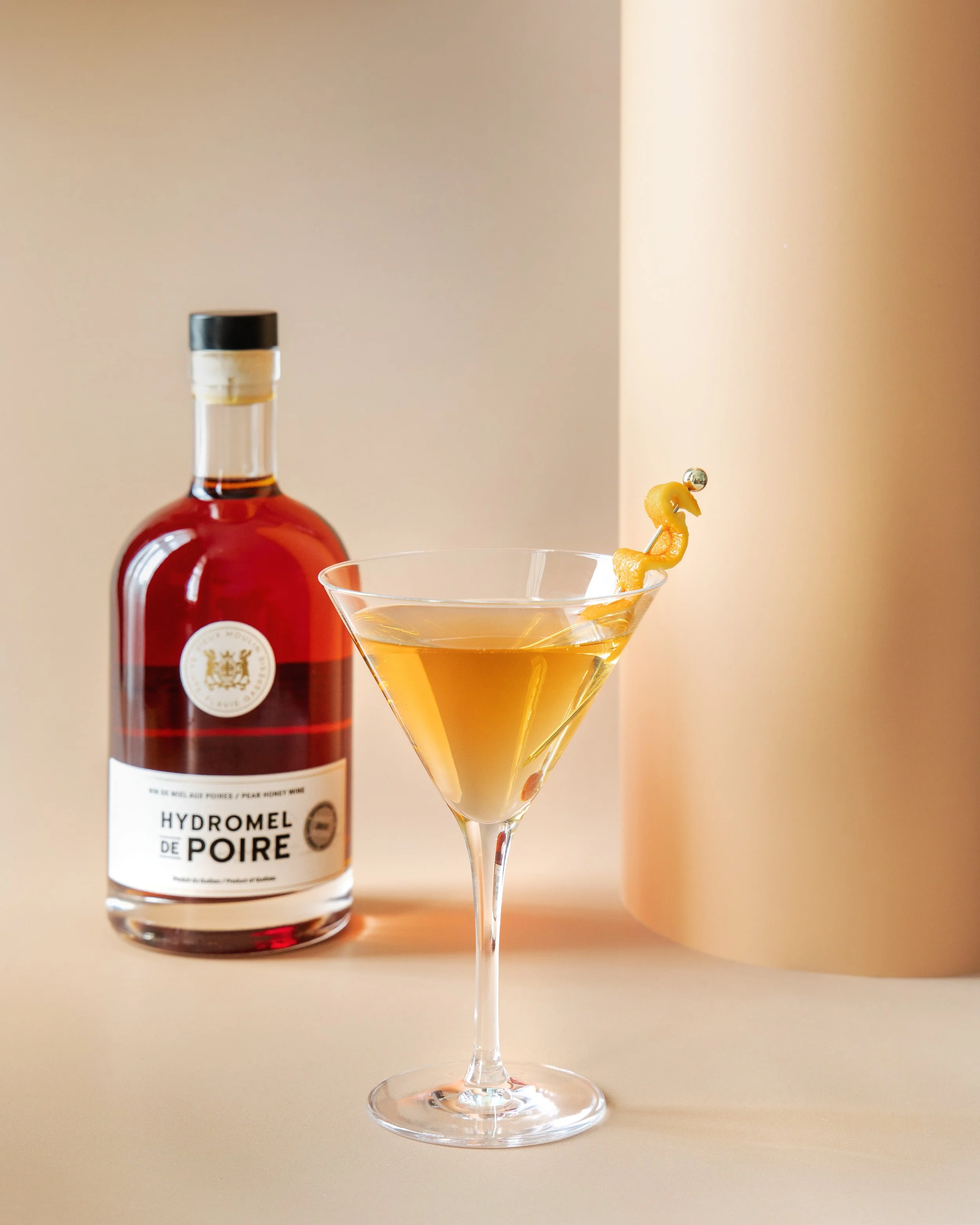 A bottle of Hydromel de Poire pear honey wine and a cocktail glass with a yellow beverage, garnished with a curled peel of pear.
