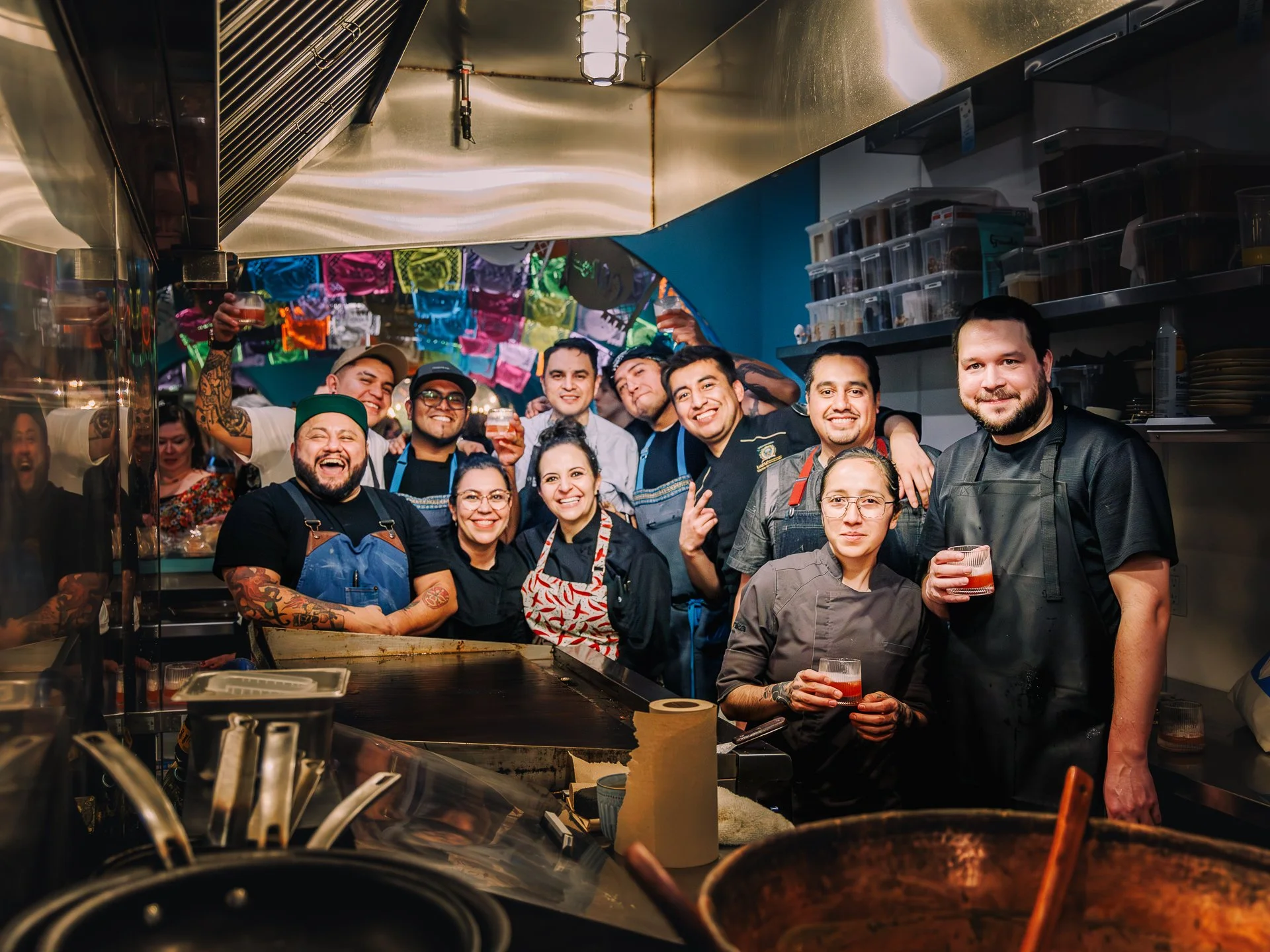 A group of diverse people smiling and holding drinks inside a restaurant kitchen with colorful decorations hanging overhead.