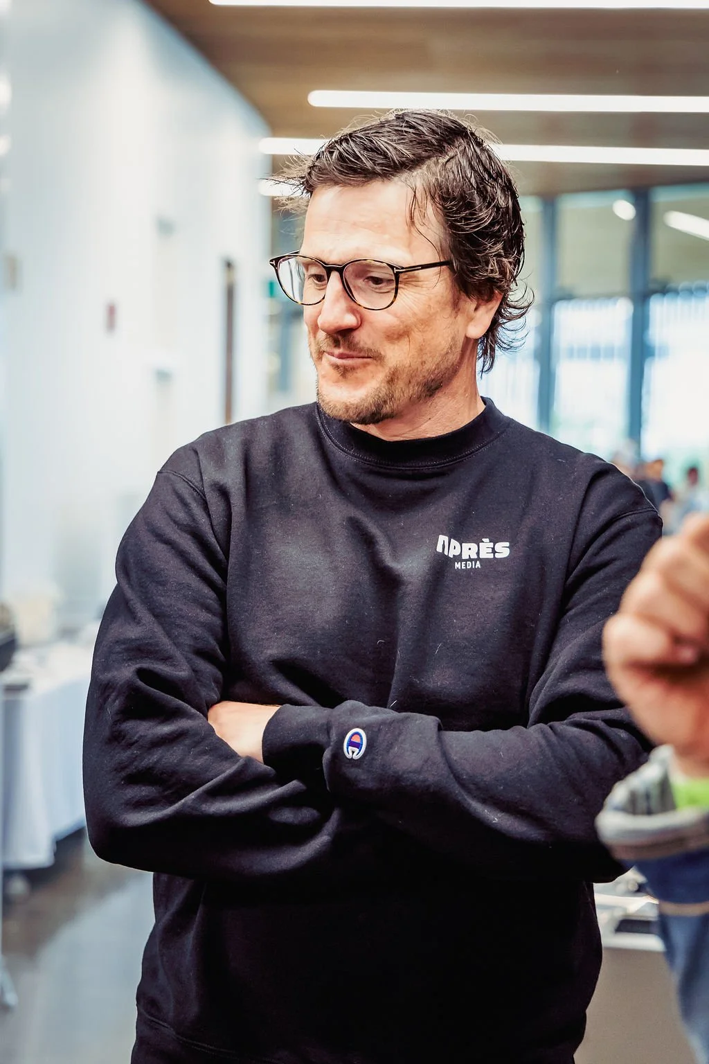 A man with glasses and dark hair, wearing a black sweatshirt with "Après Media" on it, standing with arms crossed in what appears to be an indoor space with large windows and bright lighting.