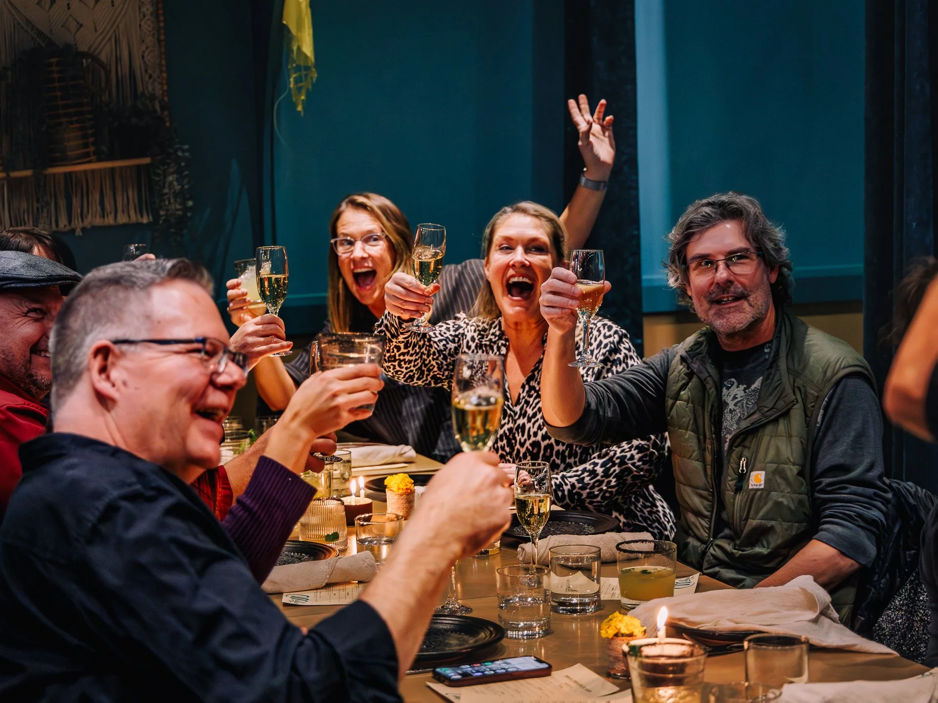 A group of people at a dinner table celebrating, holding glasses of champagne and smiling. The scene is festive with candles and a birthday cake with a lit candle on the table.