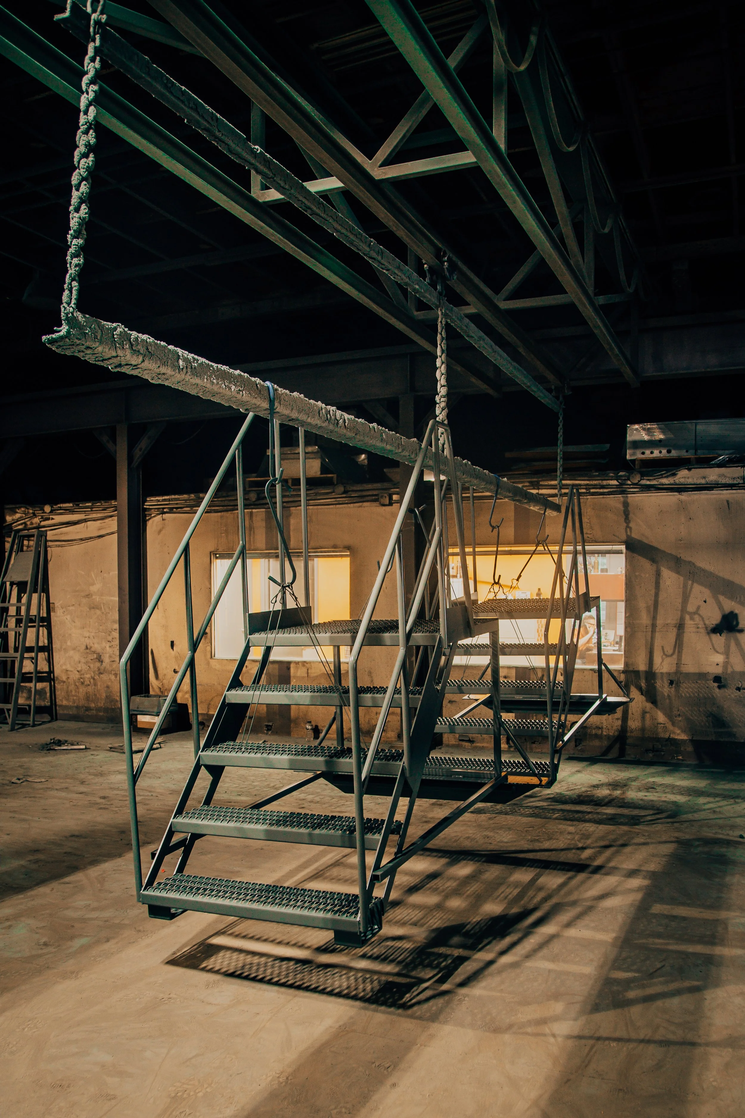 An indoor space under construction or renovation featuring a metal staircase with three steps, cast shadows on the floor, and construction materials or debris scattered around.