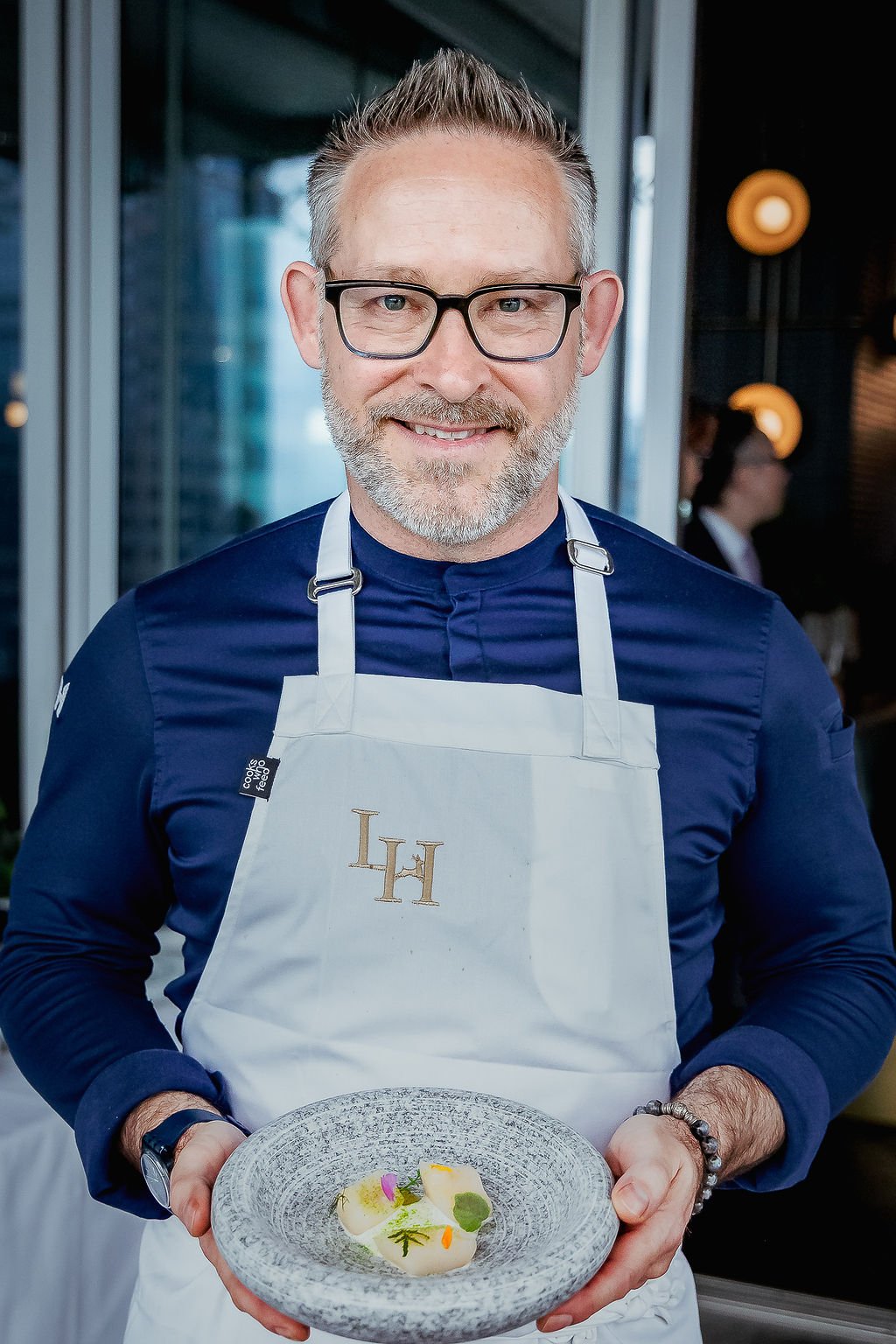A smiling man with glasses, a beard, and gray hair wearing a navy blue shirt and a white apron with embroidered initials 'LH', holding a plate with a dish garnished with herbs and edible flowers, standing in a modern restaurant setting.