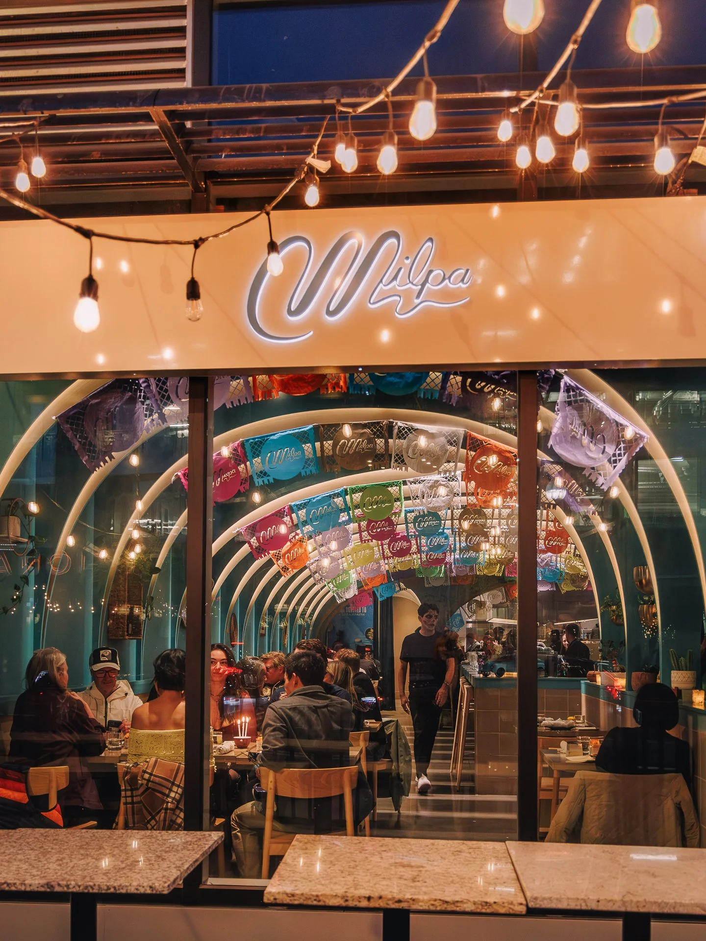 Look inside a lively restaurant with colorful Papel Picado banners, string lights, and people dining and socializing.