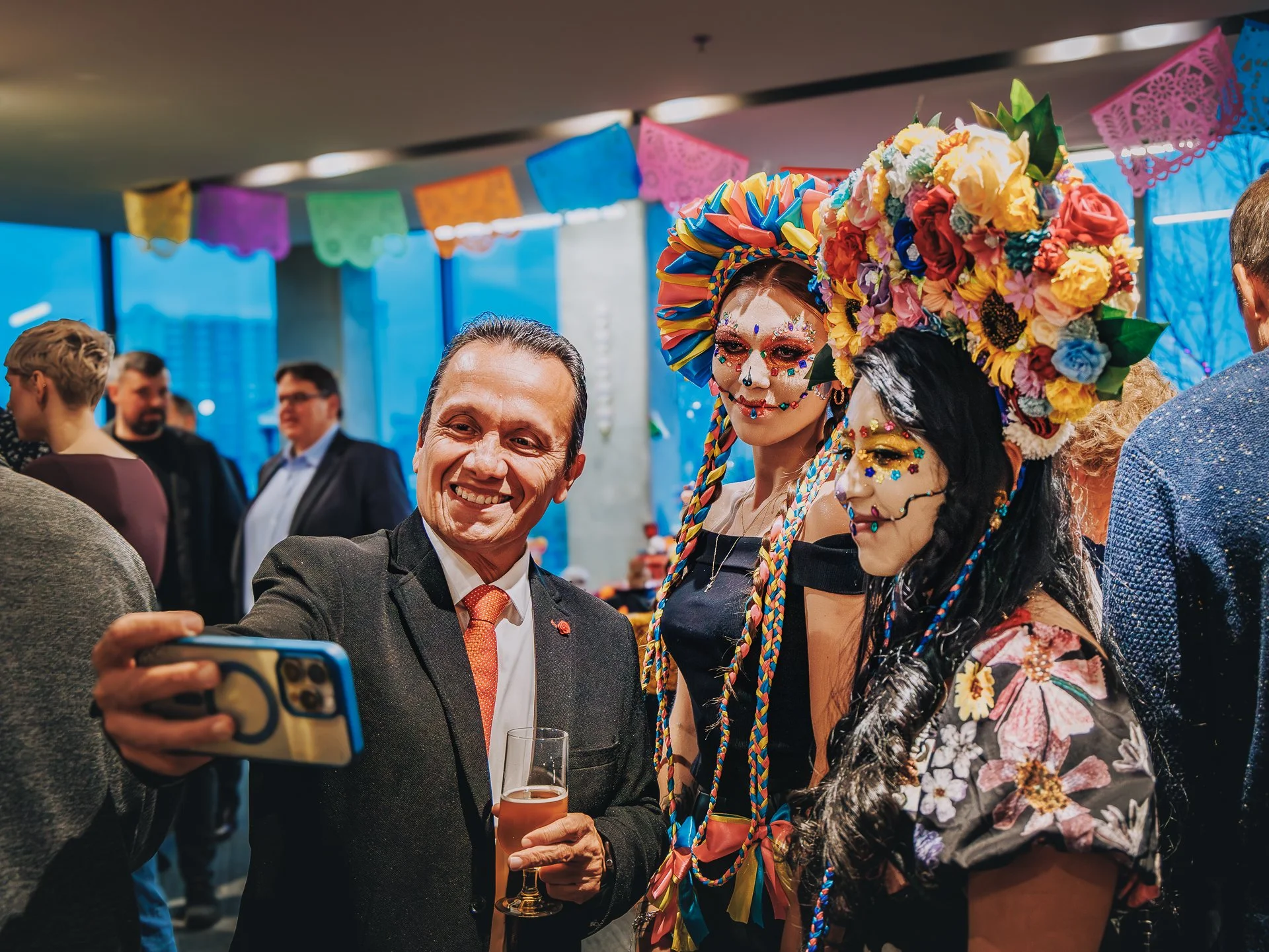 Group of people at a celebration with Mexican themed decorations, including colorful papel picado banners. One man in a suit and smiling, holding a glass of beer, is taking a selfie with a woman dressed as a La Catrina, with face makeup and a large f