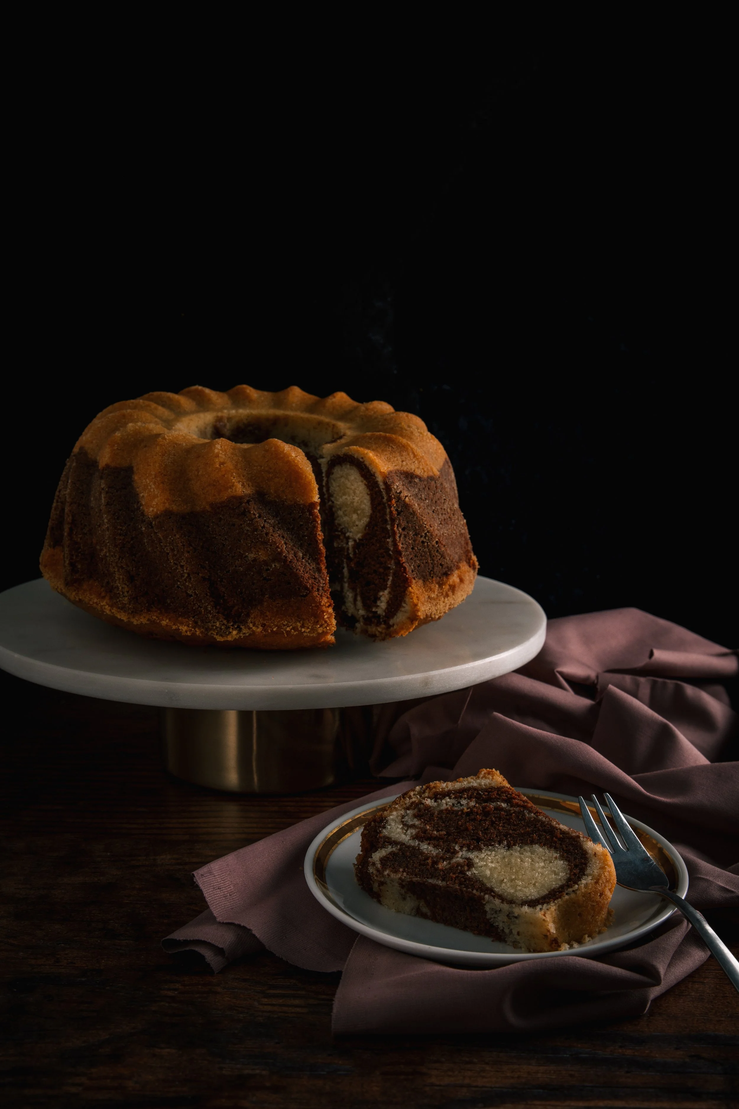 A marble Bundt cake with chocolate and vanilla swirls on a white cake stand, with a slice cut showing the marbled interior, set on a dark wooden surface with a pink napkin and fork nearby.