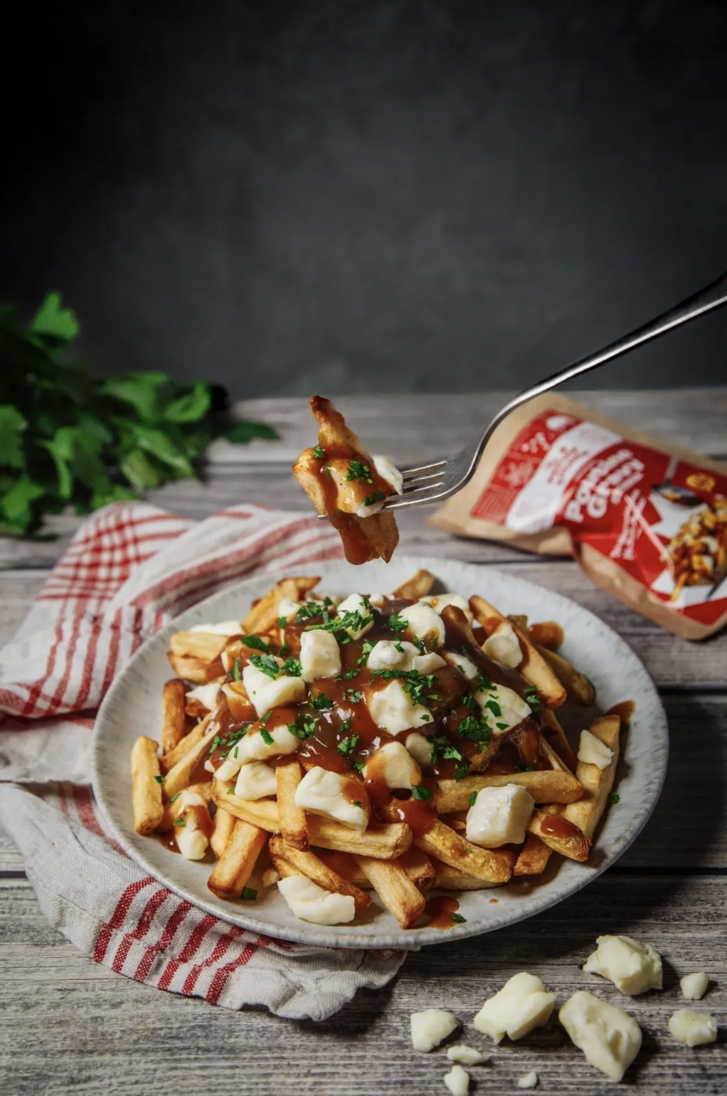 Plate of loaded fries topped with cheese, gravy, and herbs, with a fork holding a piece of fried chicken over it. There is a bag of gravy mix in the background and fresh herbs on the side, on a rustic wooden table.