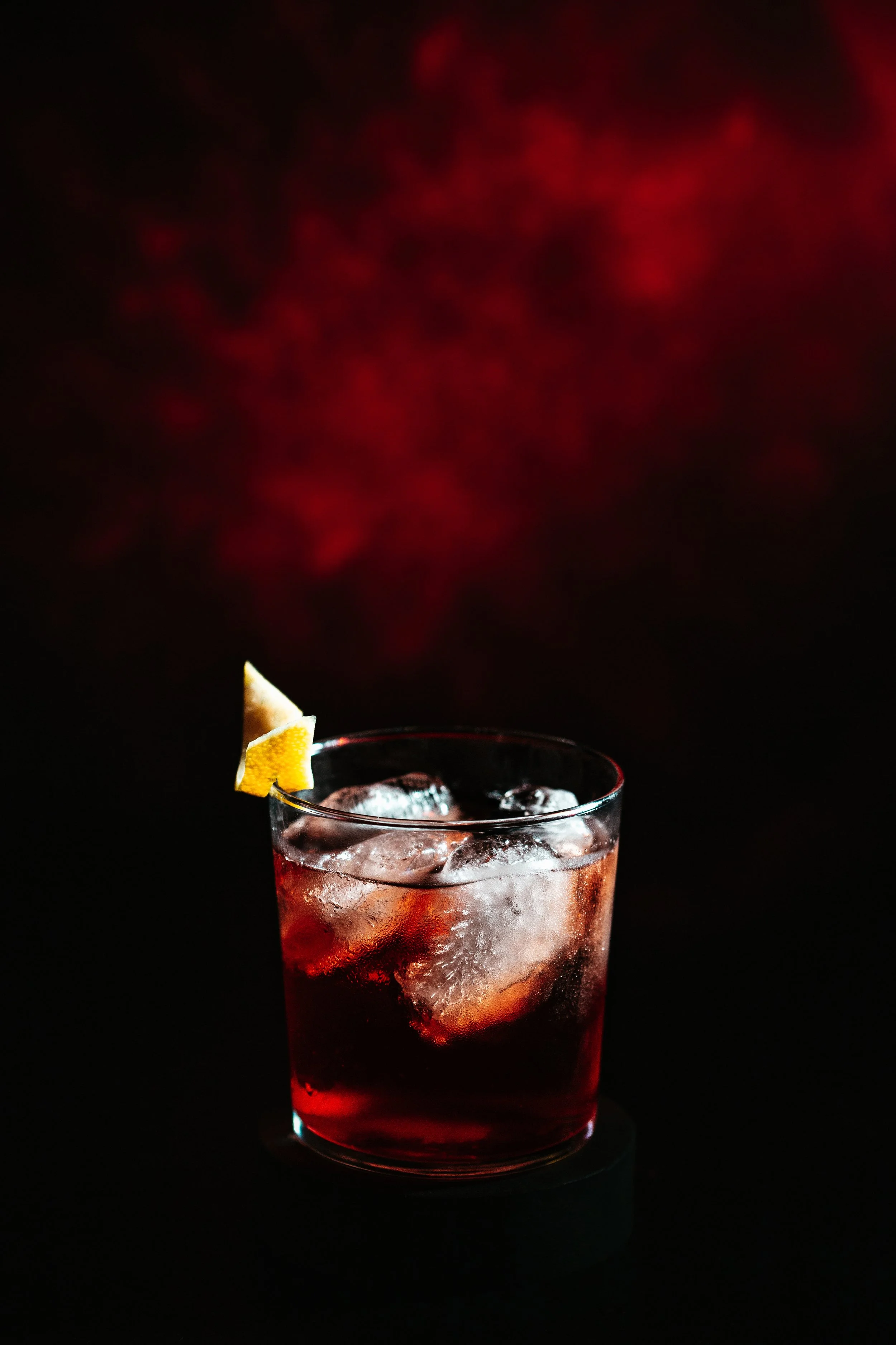 A close-up of a tall glass of red beverage with ice cubes, garnished with a lemon twist, against a dark background with red bokeh lights.
