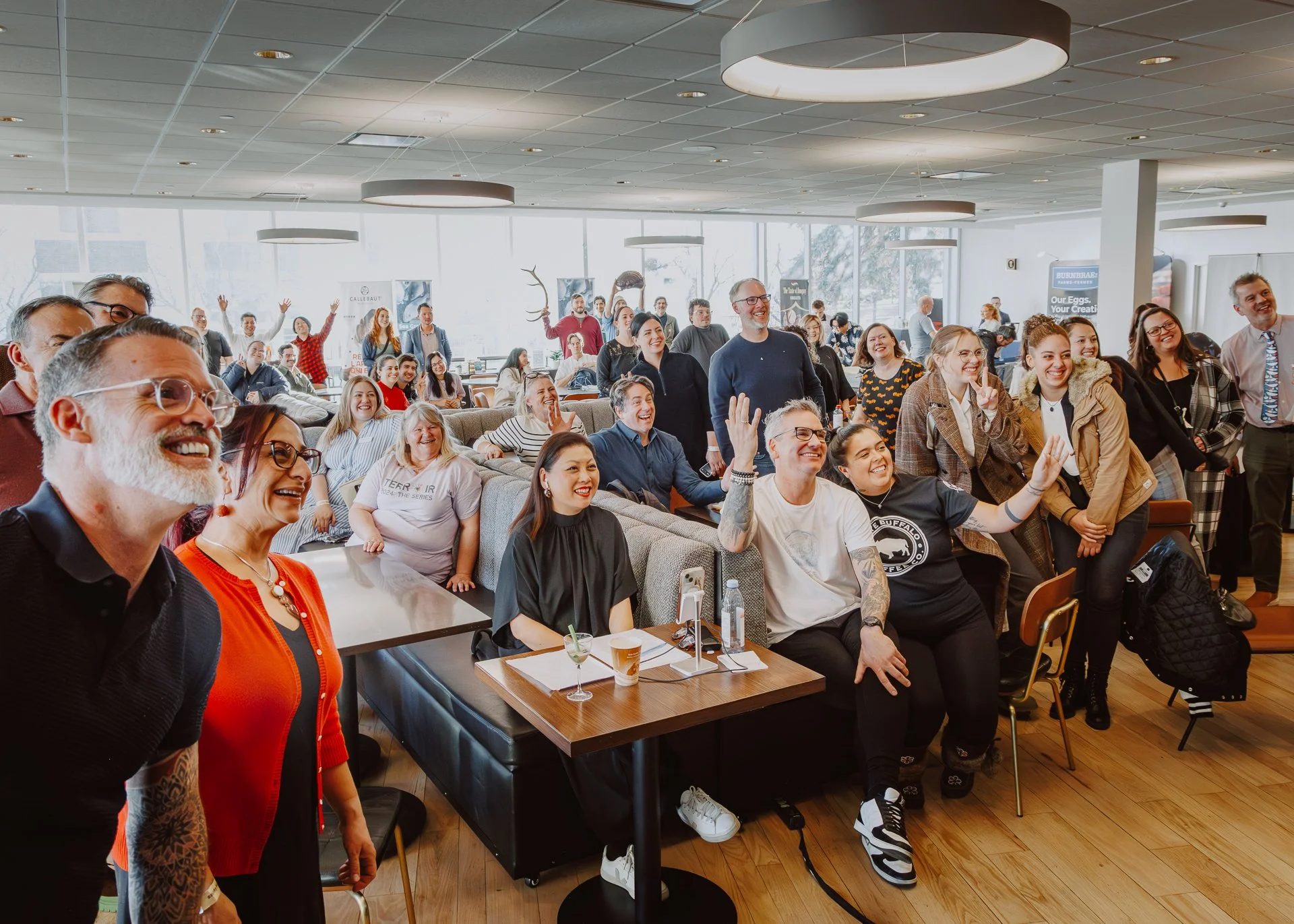A diverse group of people gathered indoors in a conference or event space, smiling and cheerfully posing for a photo, with some standing and others seated at tables.