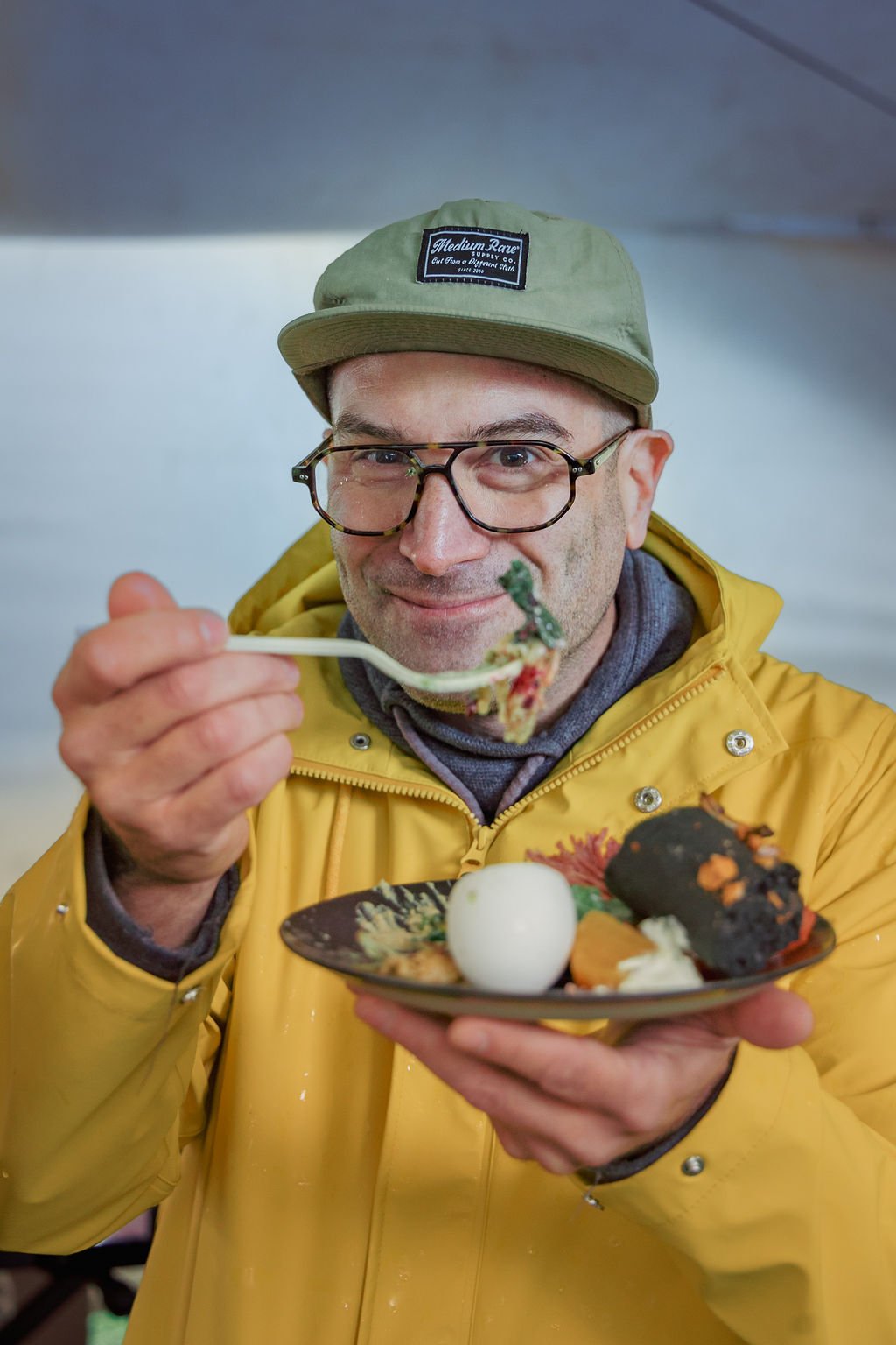 A man wearing glasses, a beige cap, and a yellow jacket is holding a plate of food with a spoon and smiling at the camera.