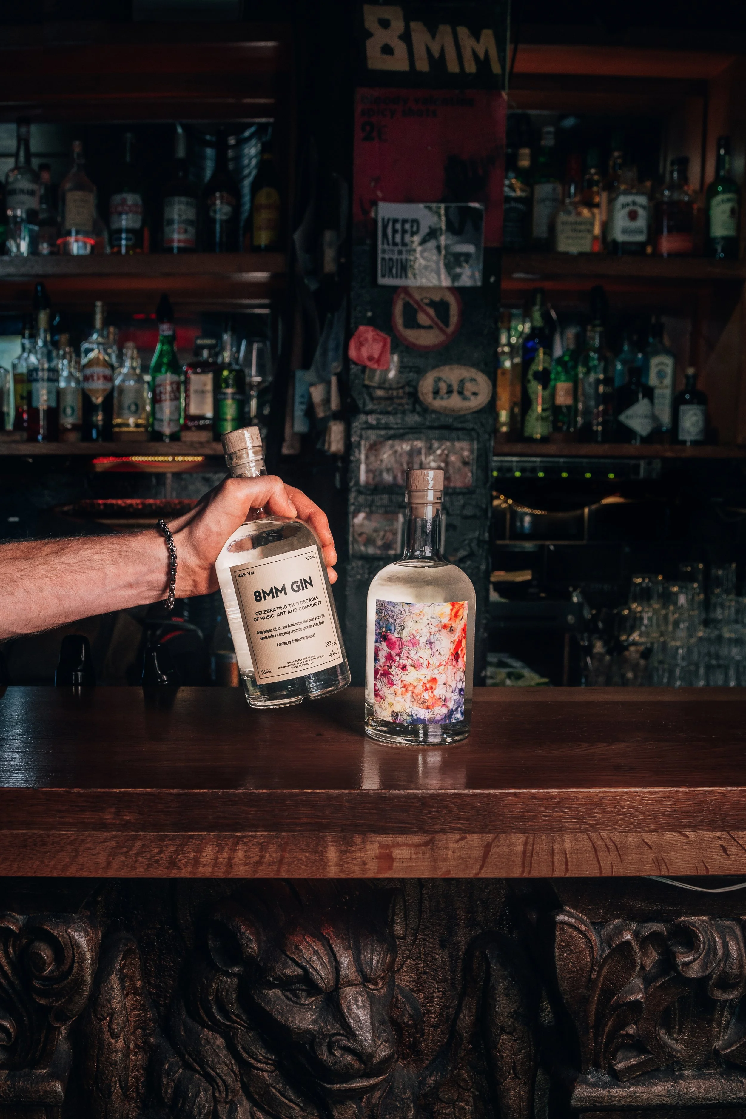 A person's hand holding a bottle of 8MM Gin in front of another bottle with colorful artwork on its label, on a wooden bar counter. The background shows shelves with various liquor bottles, a poster with multiple signs, and a dark bar environment.