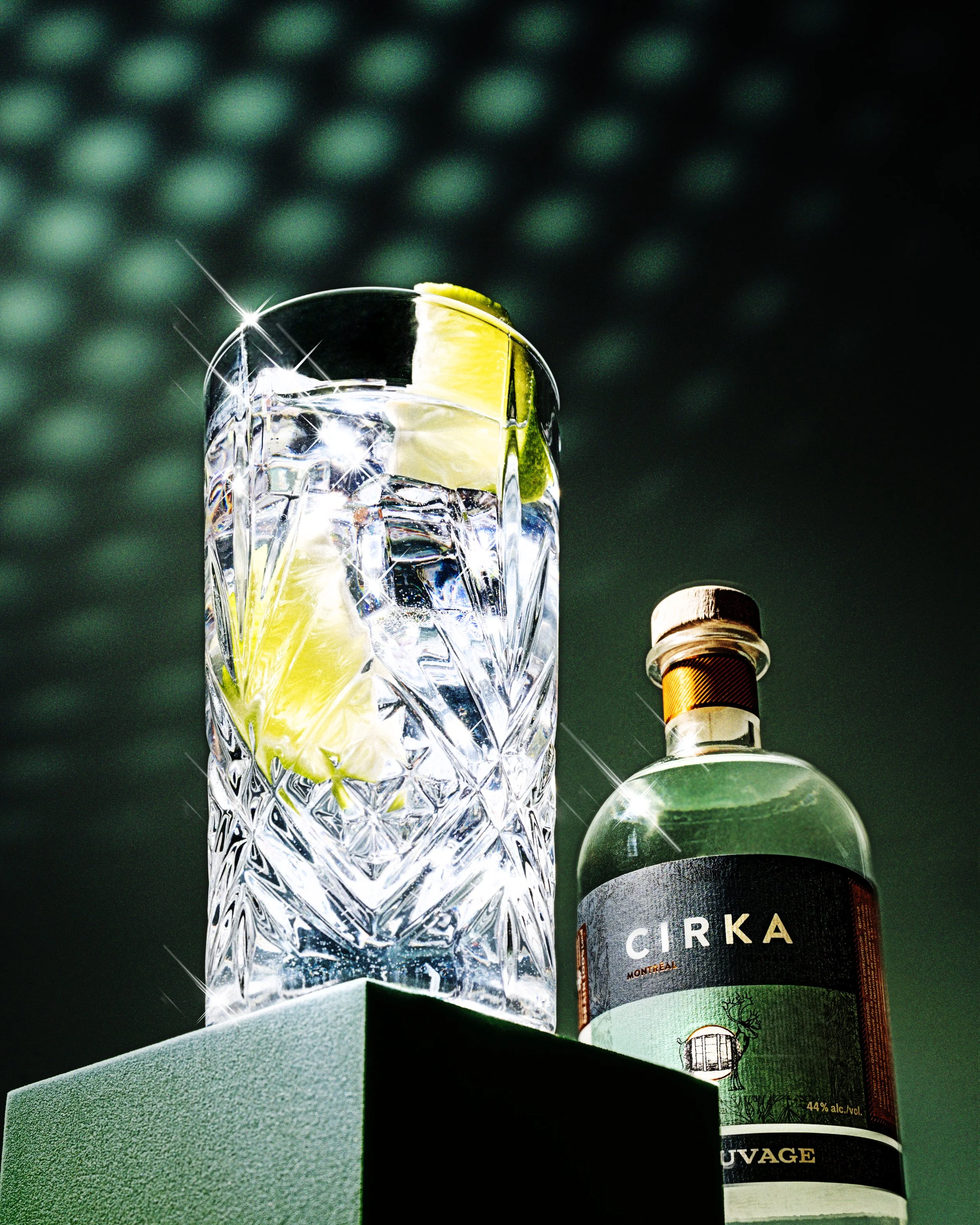 A crystal glass filled with a clear liquid and lemon wedges, illuminated by sparkling light reflections, next to a bottle of Cirka Montreal liquor with a black and white label.