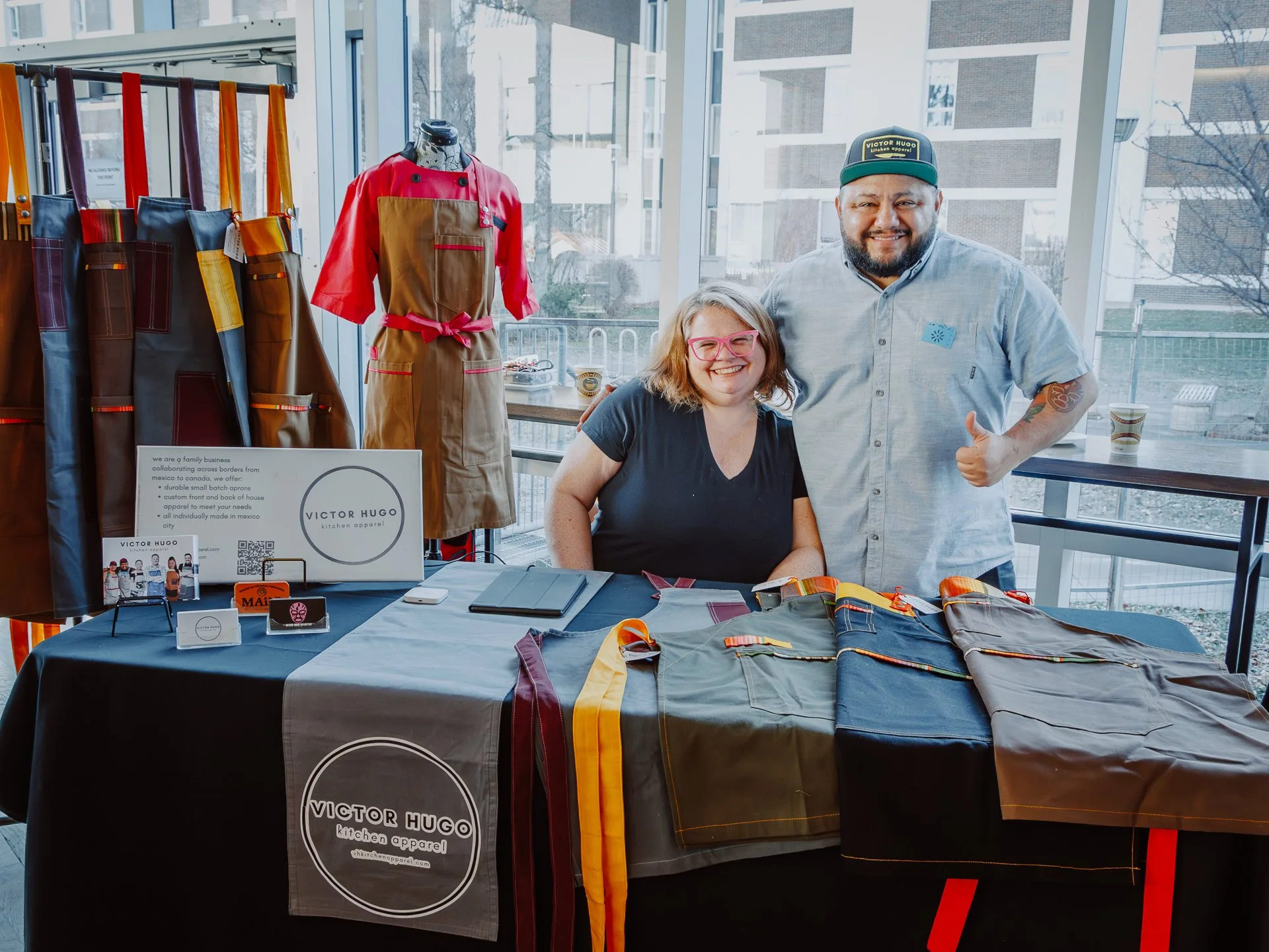 Two smiling people, a woman with pink glasses and a man with a beard, pose behind a table at a craft or vendor booth indoors with large windows. The table displays folded fabrics and a sign for Victor Hugo kitchen apparel. There are colorful aprons h