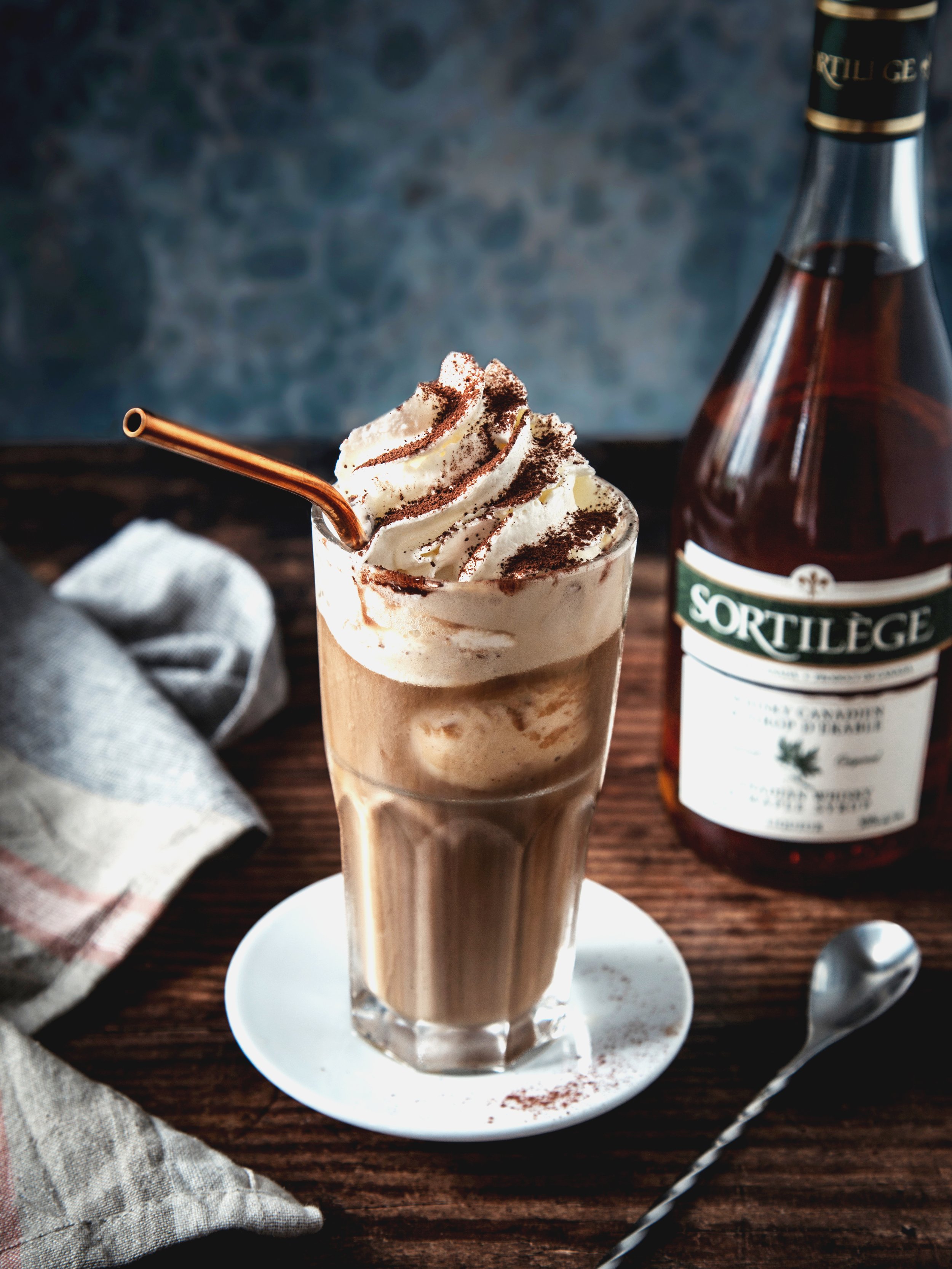 A tall glass of iced coffee topped with whipped cream and dusted with cocoa powder, with a copper straw, sitting on a white saucer on a wooden table. In the background, a bottle of Sorteige syrup and a grey cloth napkin are visible.