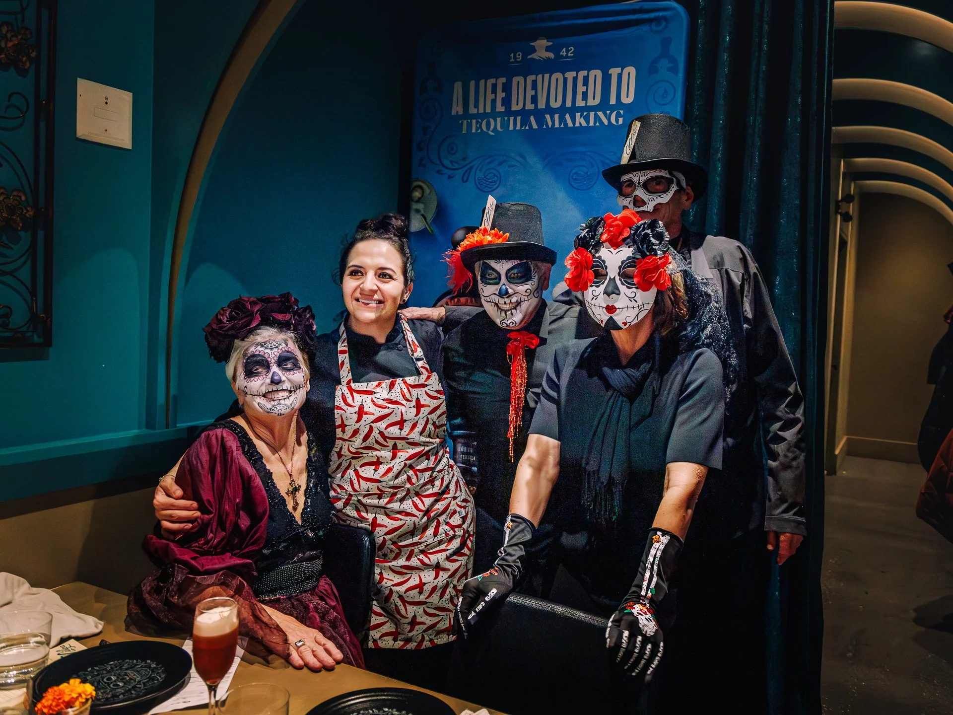 Group of people dressed in Day of the Dead costumes, including painted faces with skeleton designs and floral accessories, posing in a restaurant with a sign that says 'A life devoted to tequila making' in the background.