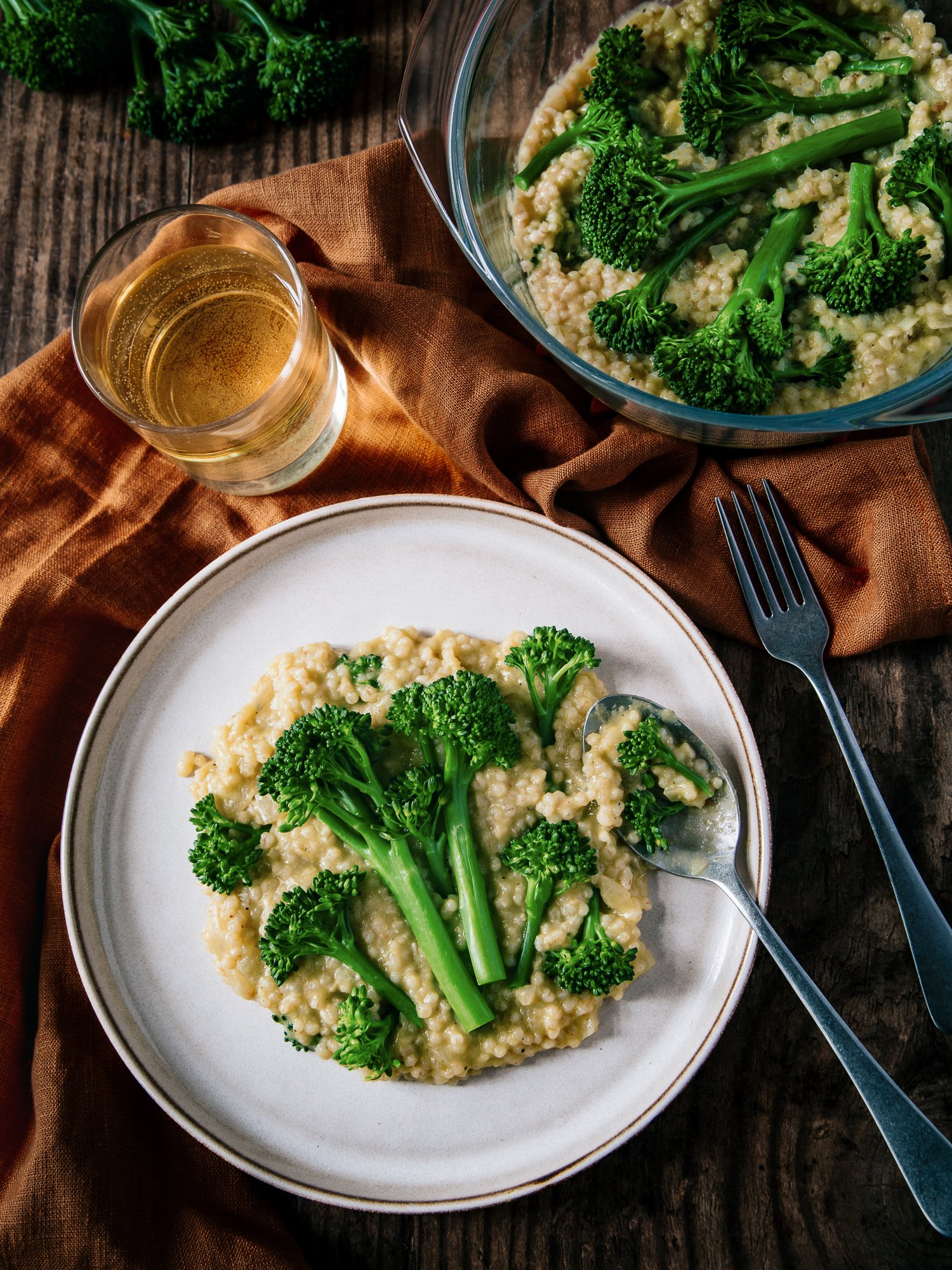 A white bowl of creamy risotto topped with fresh broccoli florets, accompanied by a fork. There is also a glass of white wine, and a larger glass bowl of more broccoli and risotto on a rustic wooden table, with a brown napkin underneath.