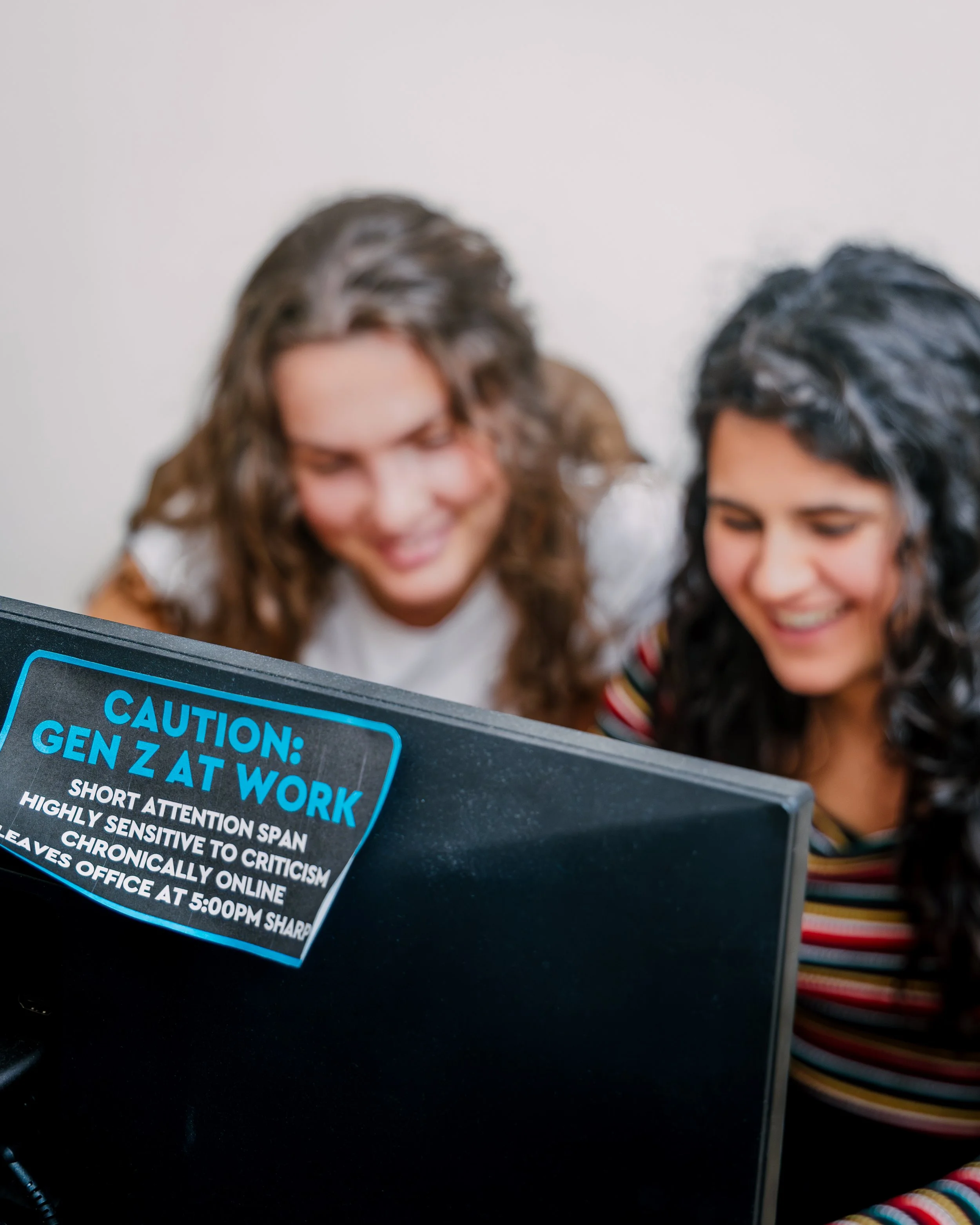 Two women with hair covered in shampoo or conditioner, smiling while looking at a device on a black surface with a caution sign that warns about gen Z at work.