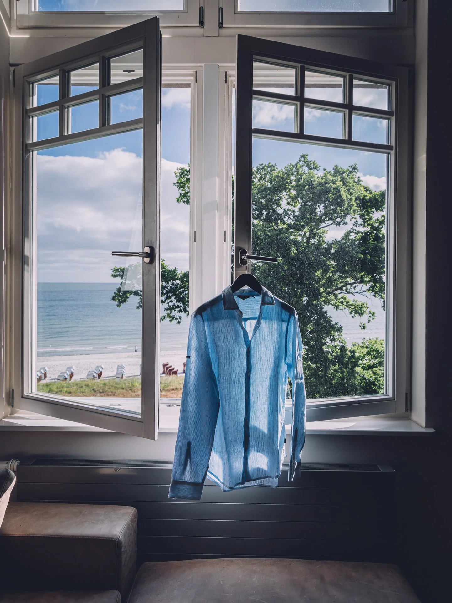 Open window with a view of a beach, trees, and ocean, with a blue shirt hanging on the window latch.