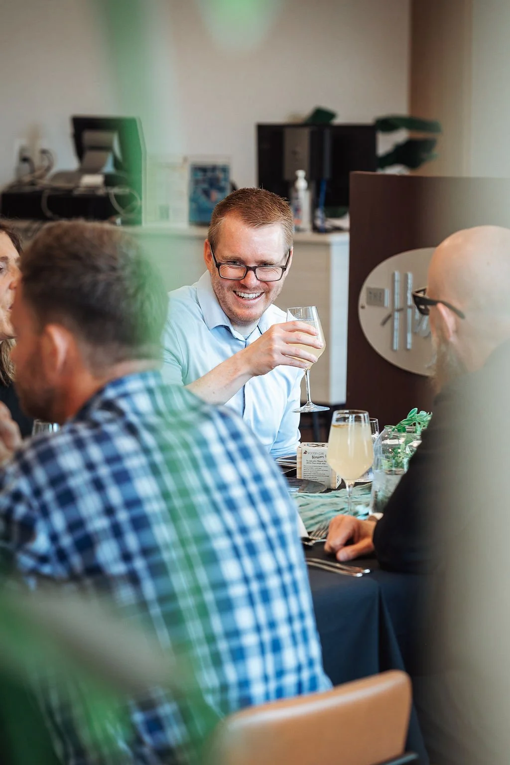 A group of people sitting at a dinner table, laughing and enjoying drinks, with a man in glasses holding a glass of white wine.