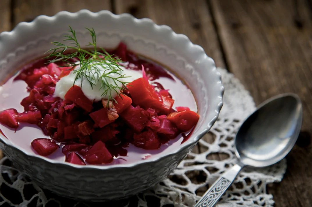 A bowl of beet soup garnished with sour cream and fresh dill, placed on a crocheted lace doily, with a silver spoon on the side.