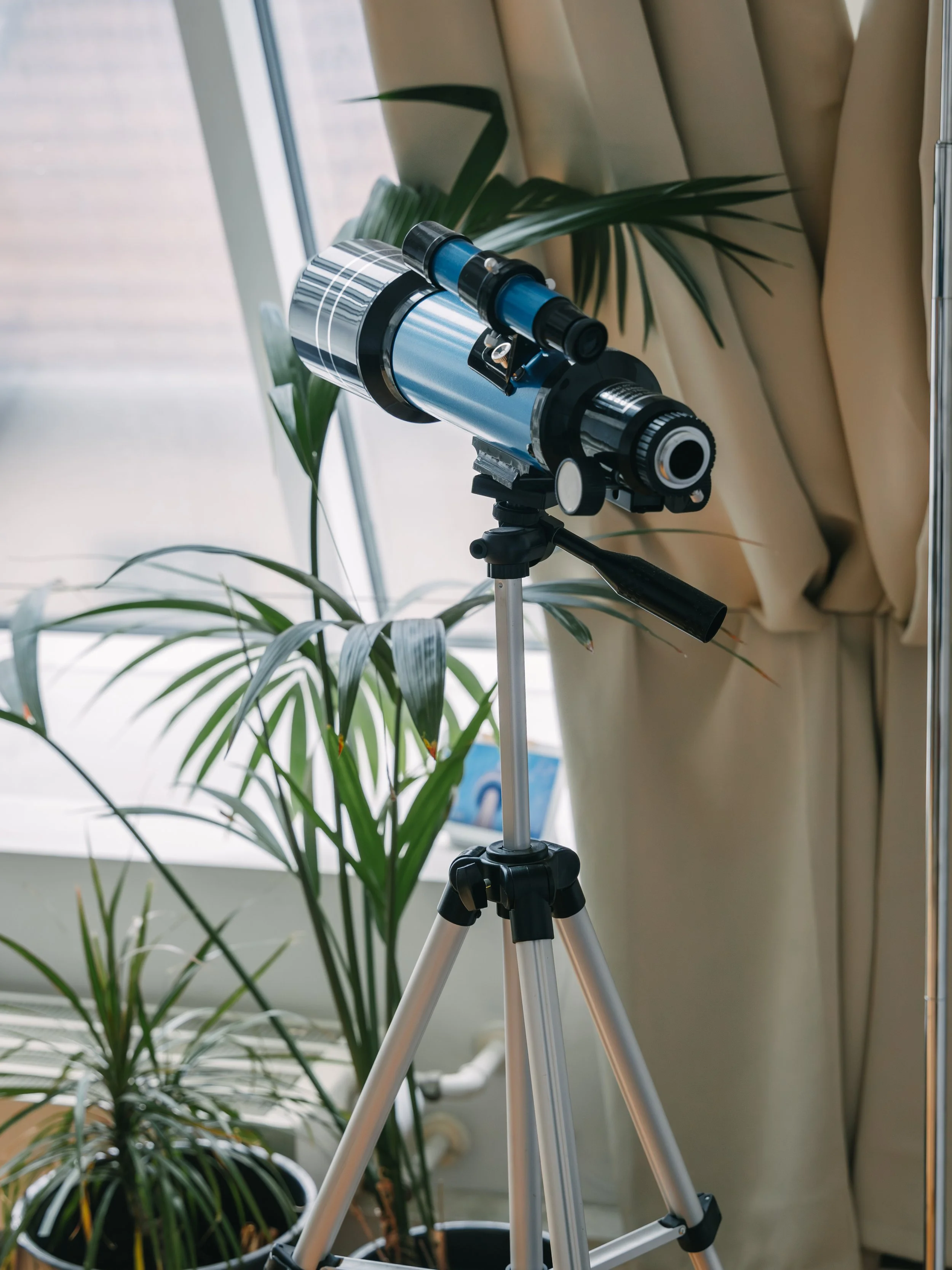 A blue and black telescope on a tripod, positioned indoors near a window with green houseplants nearby.