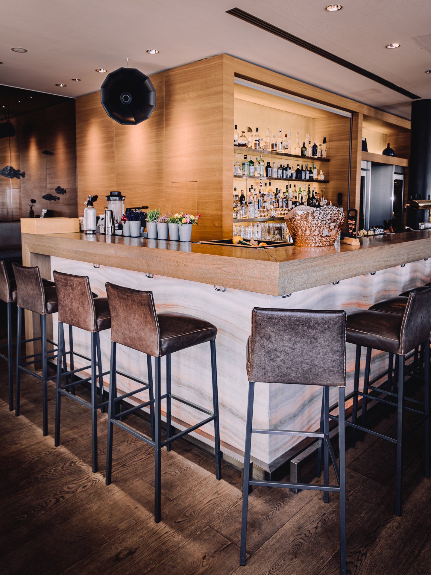 A bar with a wooden counter and high chairs made of metal and brown leather, a marble base, and a backdrop of shelves with assorted liquor bottles, a basket, flowers, and a black chalkboard sign.