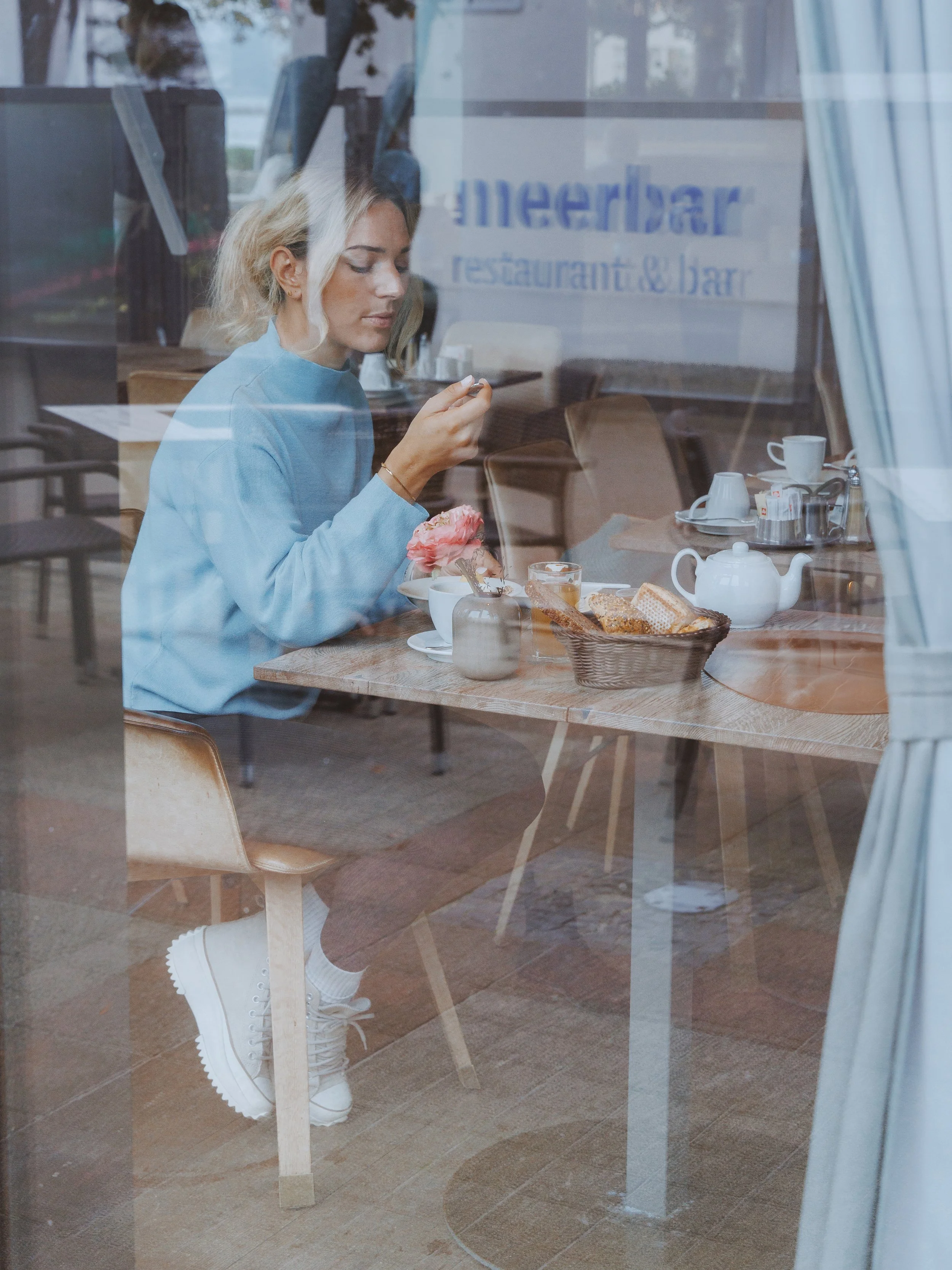 A woman with blonde hair wearing a light blue shirt and white sneakers sitting at a table in a cafe, looking at a smartphone, with dessert and tea on the table, seen through a glass window.