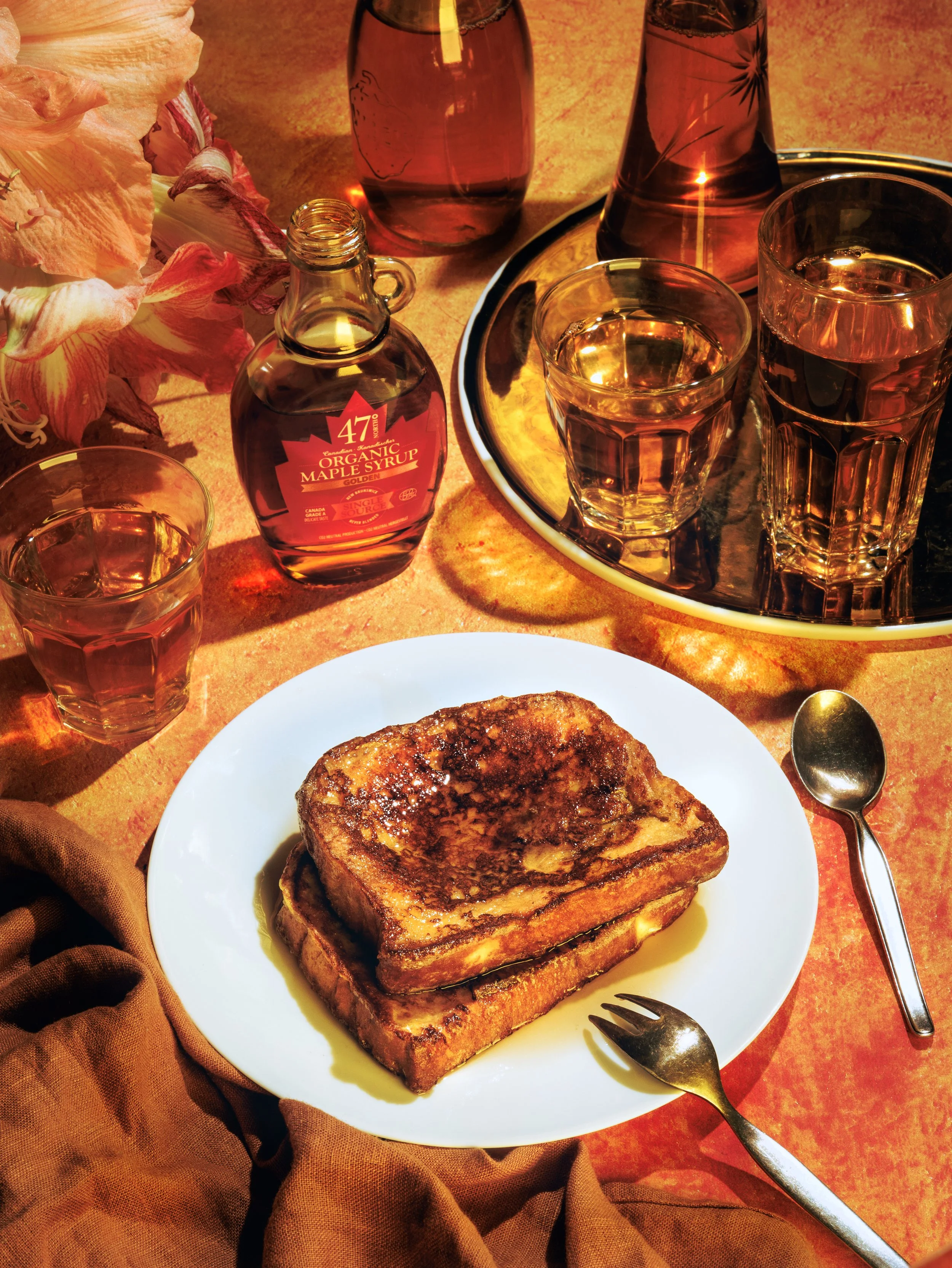 A plate with toasted bread, a glass of water, and a small bottle of maple syrup on a dining table with a bunch of pink flowers and a tray with glasses and bottles in the background.