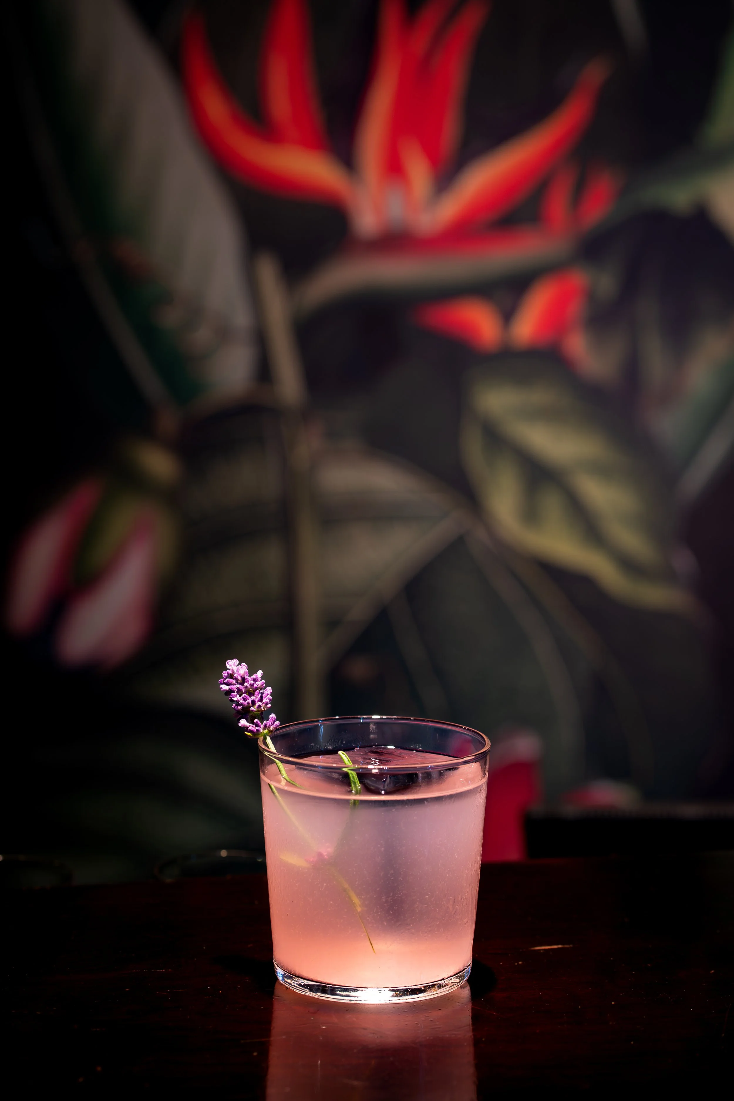 A pink-colored beverage in a clear glass with a sprig of lavender resting on the rim, set on a dark wooden surface with a blurred background of tropical or garden plants.