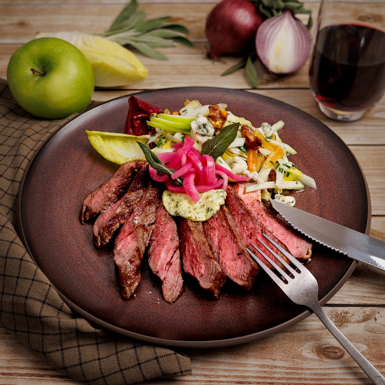 A plate with sliced cooked steak, topped with pickled red onions, served with salad and garnished with herbs, set on a wooden table with a glass of red wine, a green apple, pineapple slice, and red onion in the background.