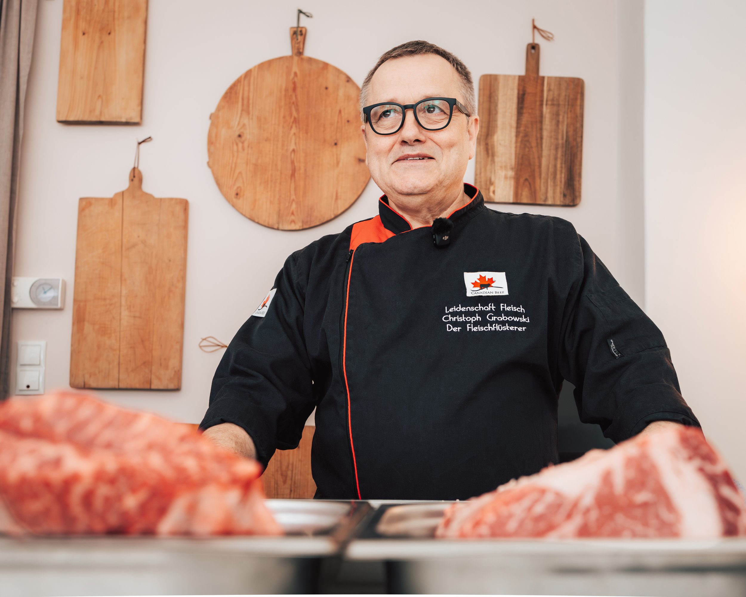 A man with glasses wearing a black chef's jacket with text on it stands behind a counter, looking to the side, with wooden kitchen tools hanging on the wall behind him.