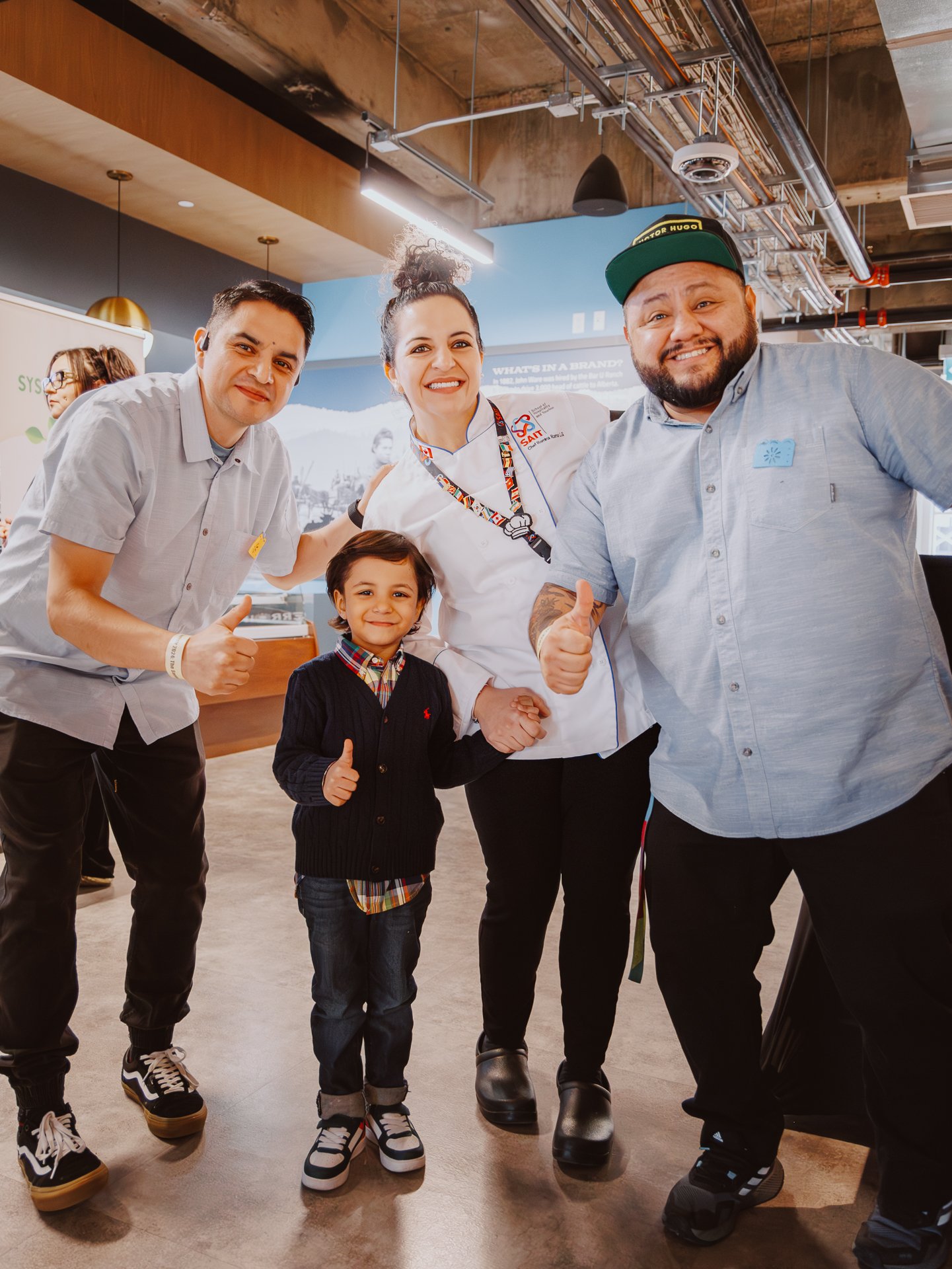 Group of four people, including a young boy, smiling and giving thumbs-up, indoors with industrial ceiling, two men and one woman with two other women in the background.