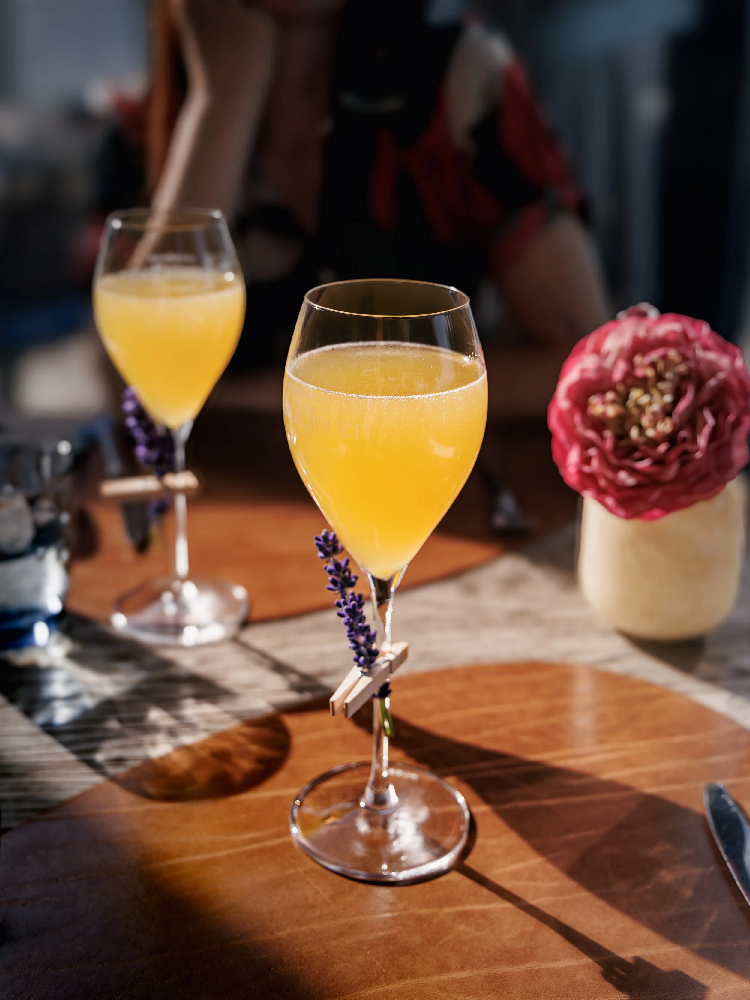 Two glasses of yellow drink on a wooden table with a person in the background and a pink floral arrangement on the side.