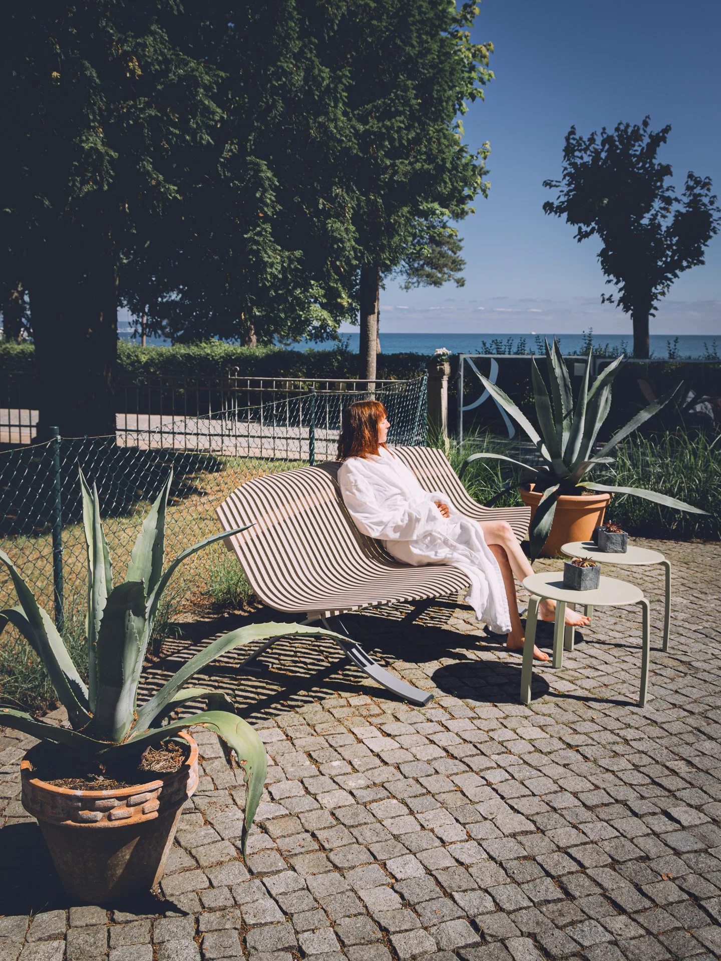 A woman in a white dress relaxing on a striped outdoor bench next to potted aloe plants, with large green trees, a fence, and a view of the ocean under a blue sky.
