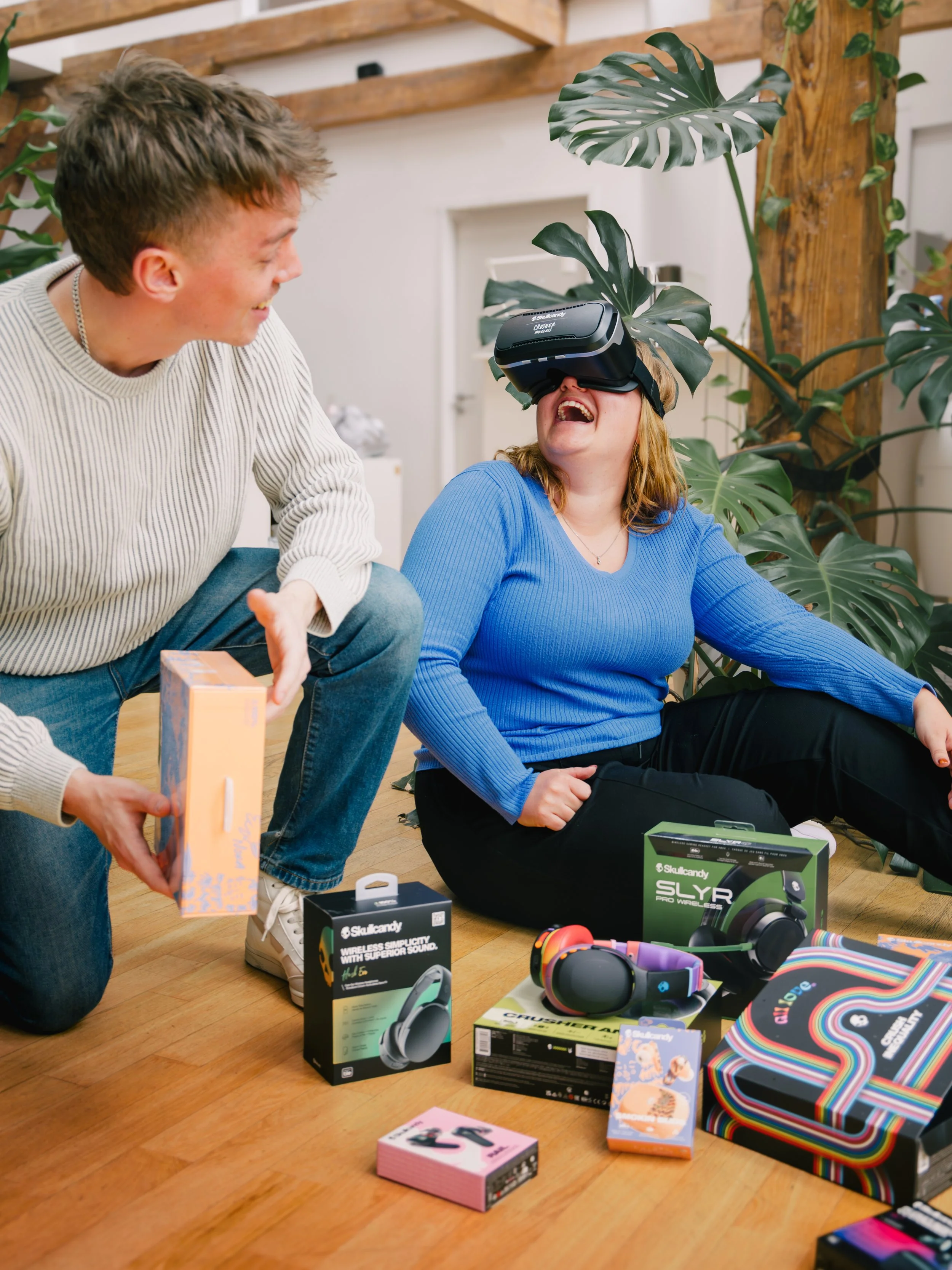 A young person wearing a virtual reality headset and a woman smiling at her with a gift box in her hand in a room decorated with large green houseplants, and various tech gadgets and boxes on a wooden floor.