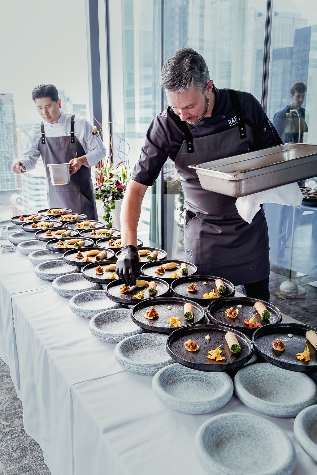 Two chefs preparing multiple styled dishes on black plates, with one placing a garnish on each dish. The setting appears to be a high-rise restaurant or banquet with city skyline view.