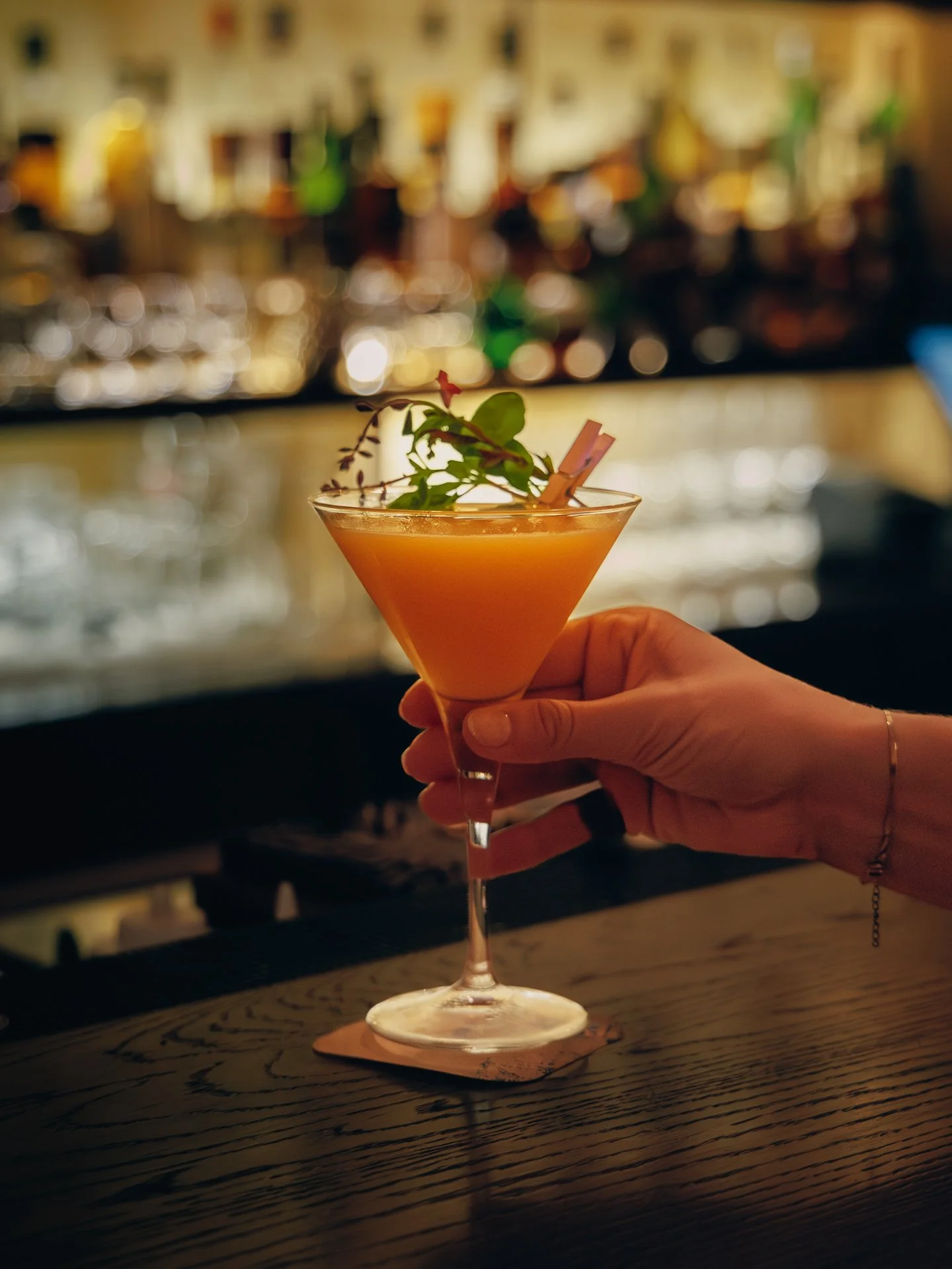 A hand holding a cocktail glass with an orange drink garnished with green herbs and a small pink umbrella, set on a wooden bar with blurred bar shelves in the background.