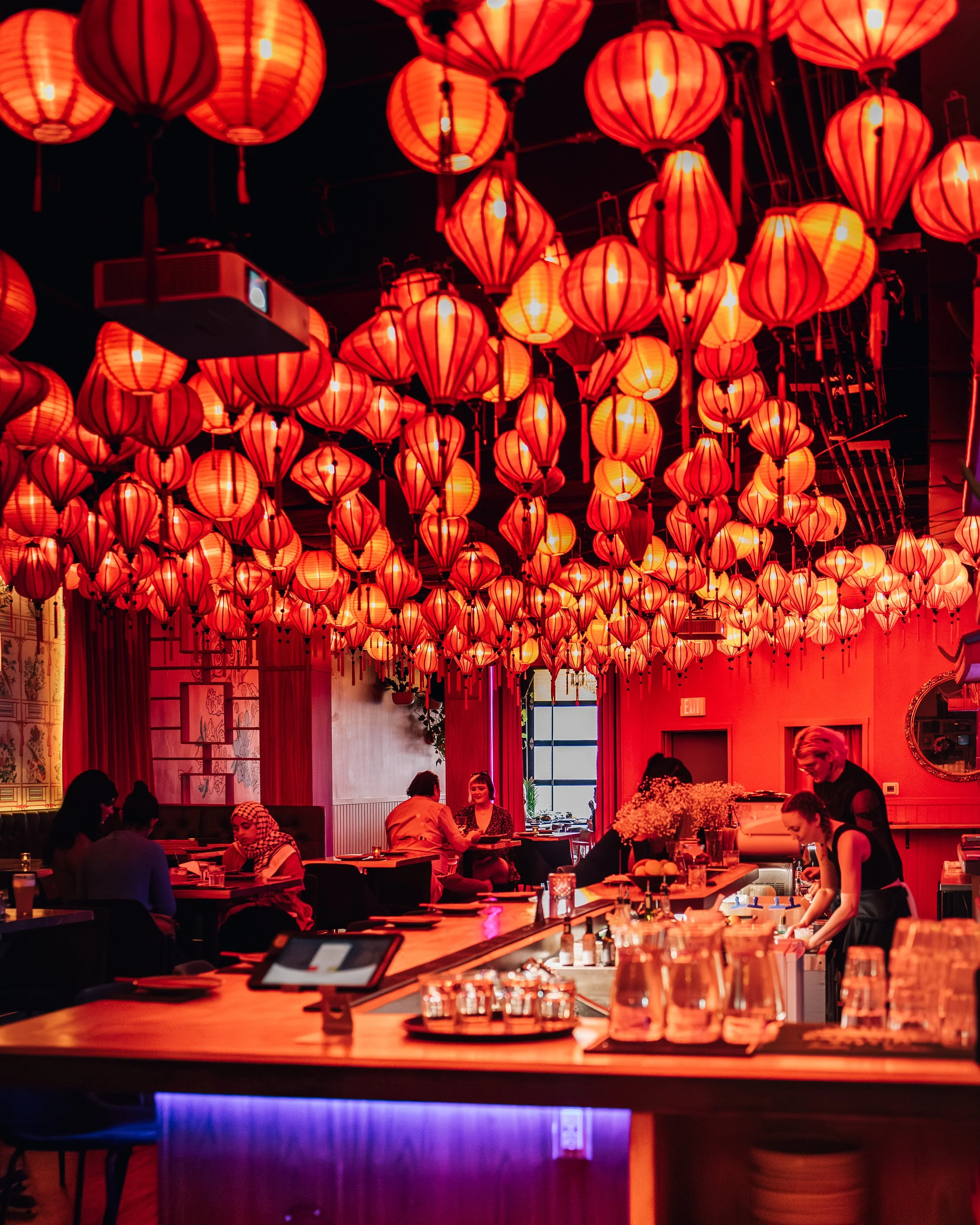 Interior of a restaurant decorated with red paper lanterns hanging from the ceiling, with people dining and a bar counter in the foreground illuminated with colorful lights.