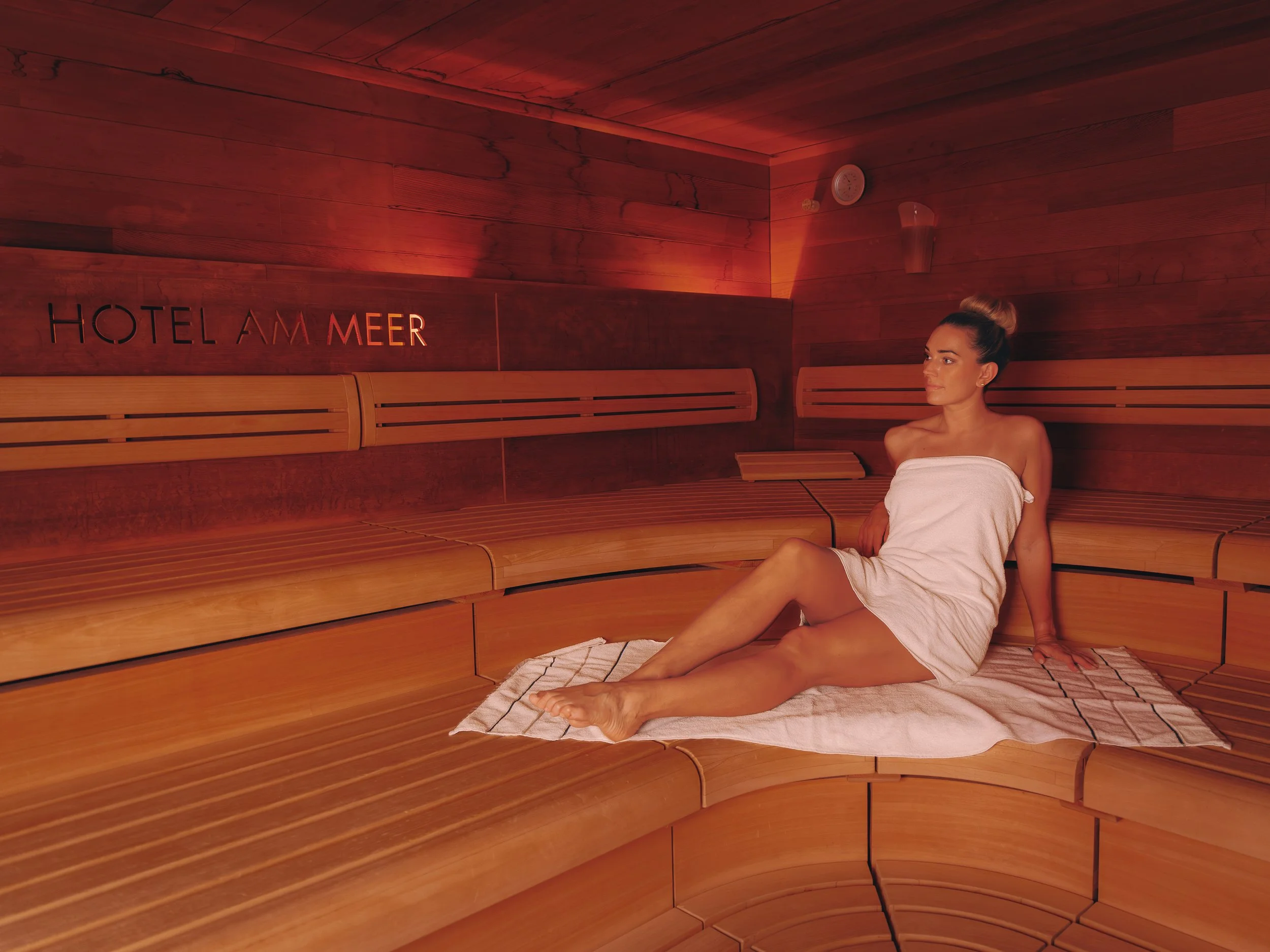 A woman sitting inside a wooden sauna, wrapped in a towel, with a relaxed expression. The sauna has wooden benches and walls, with a wall-mounted thermometer and a light fixture visible. The sign on the wall reads 'HOTEL AM MEER'.