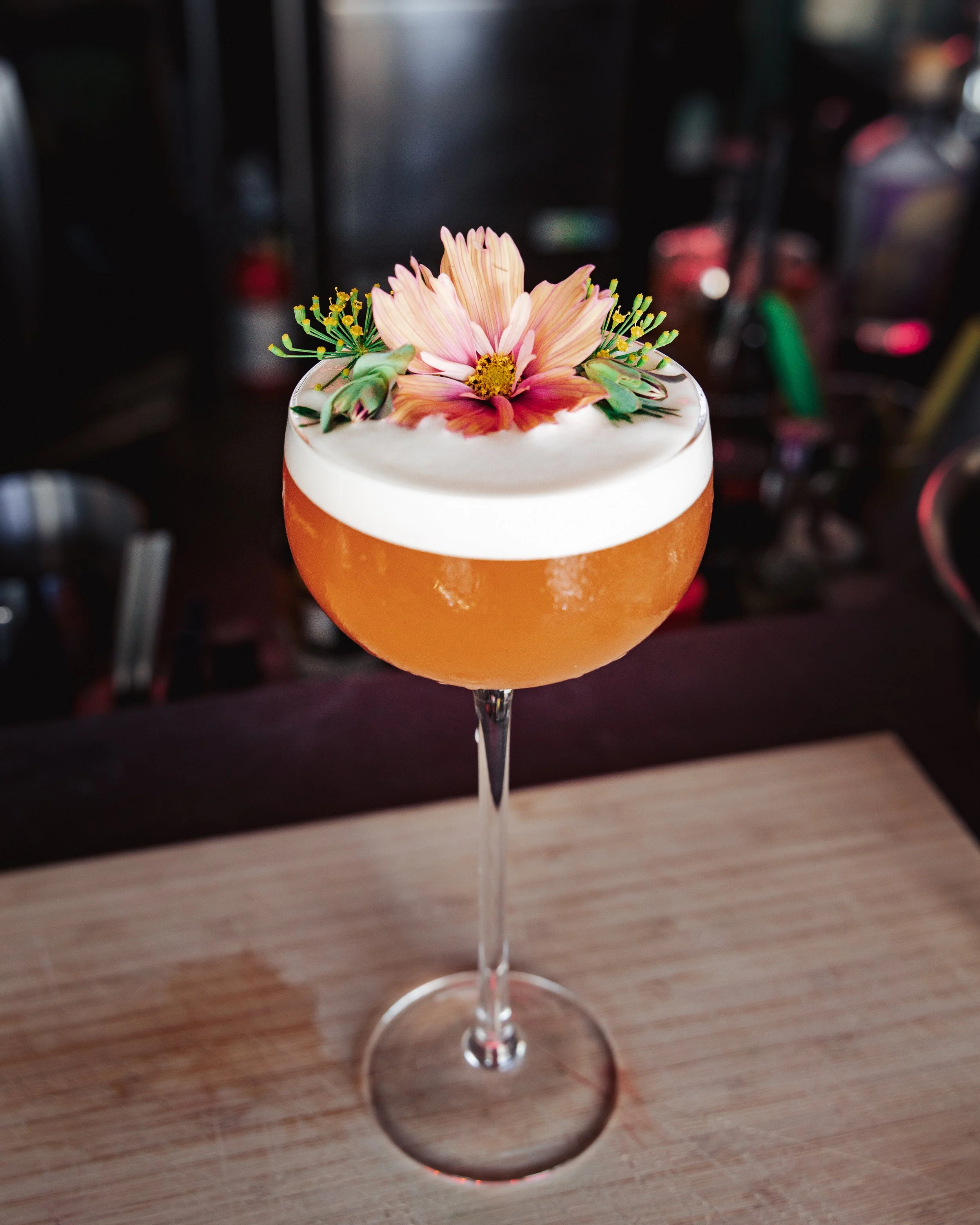 A cocktail glass filled with an orange-colored drink, garnished with a pink flower, green decorative picks, and a small yellow flower, placed on a wooden surface.