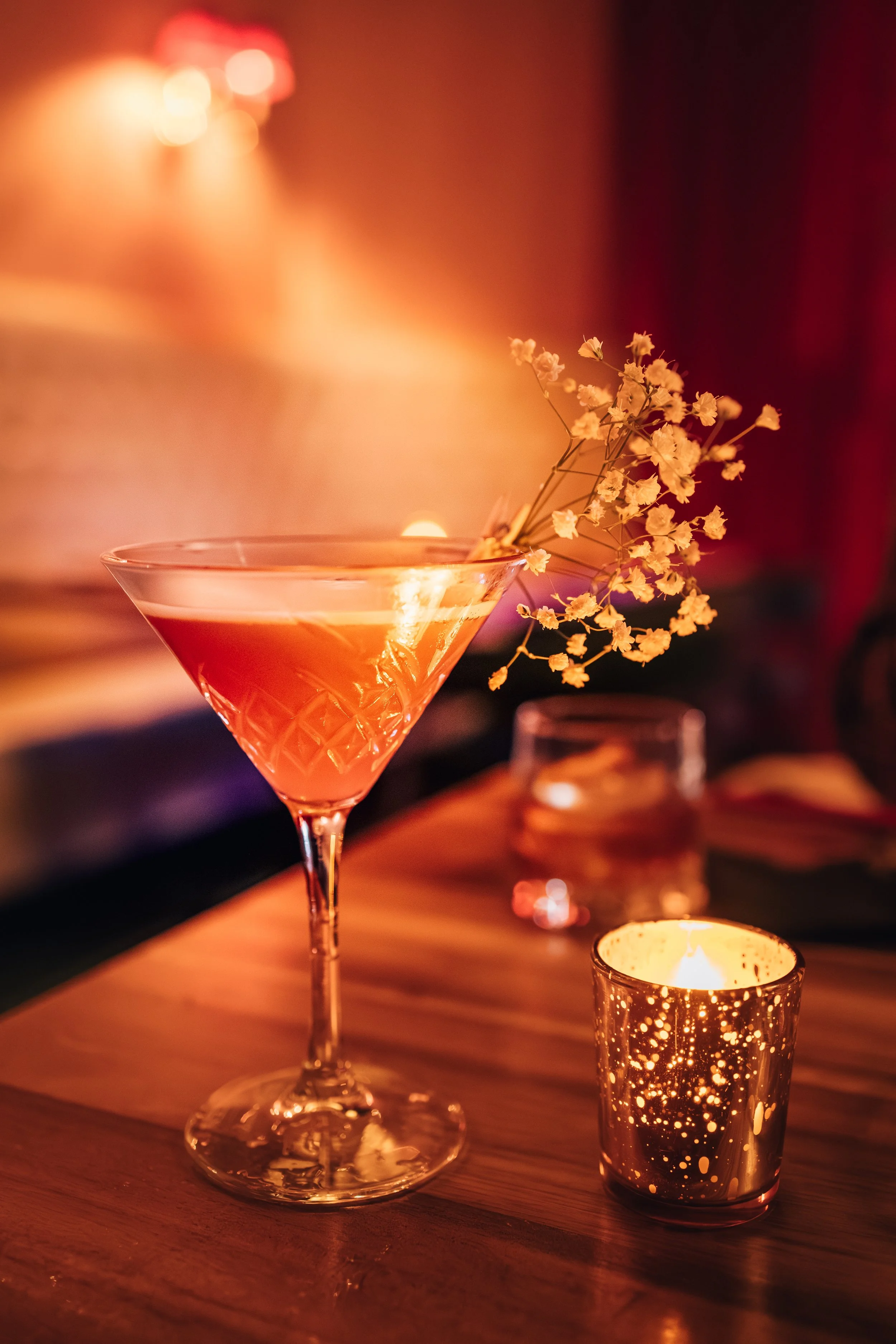 A cocktail glass with a pink drink garnished with small white flowers, set on a wooden table with a lit candle in a decorative holder nearby, in a dimly lit setting.
