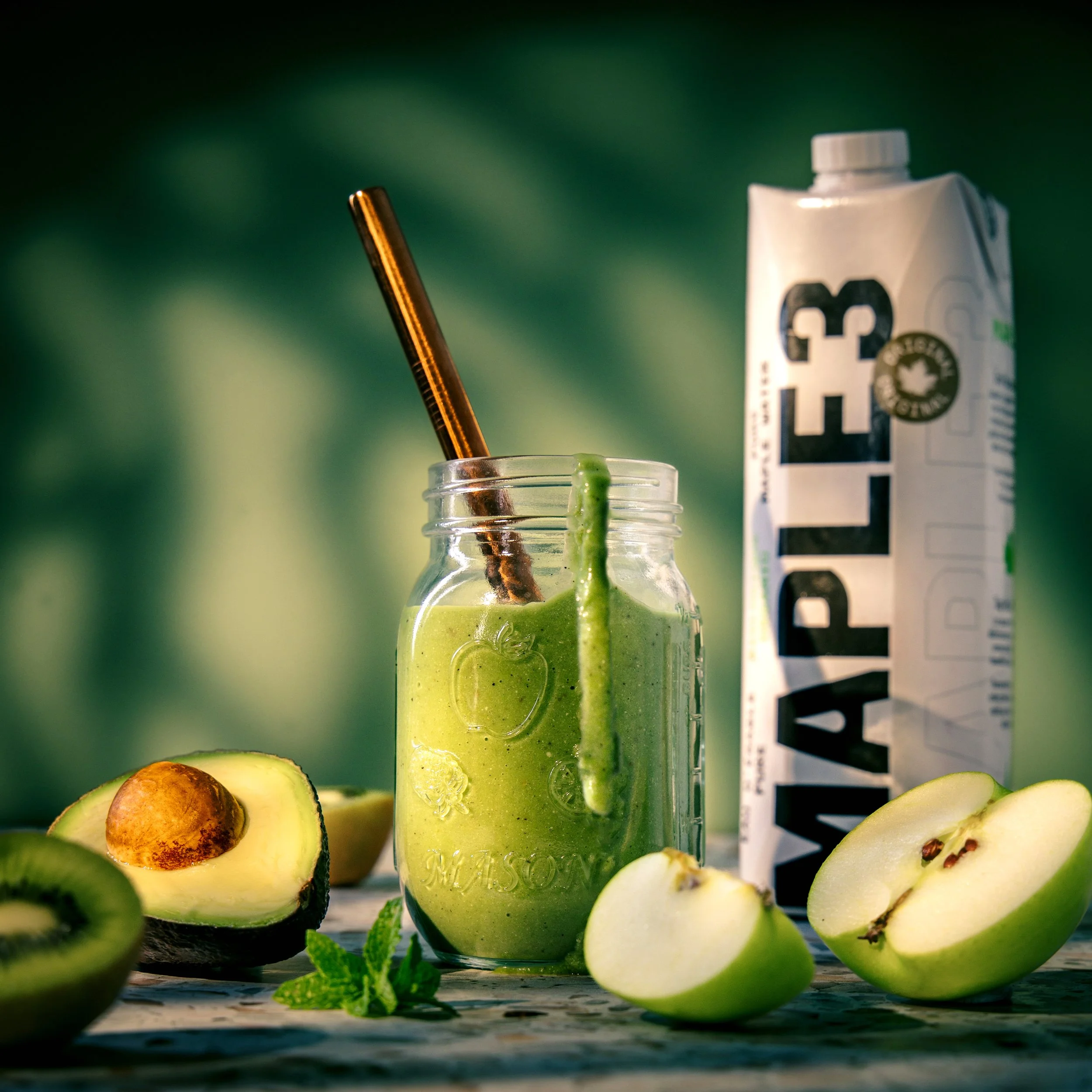 Close-up of a blender filled with green smoothie, surrounded by halved avocados, mint leaves, and a carton of plant-based milk in the background.