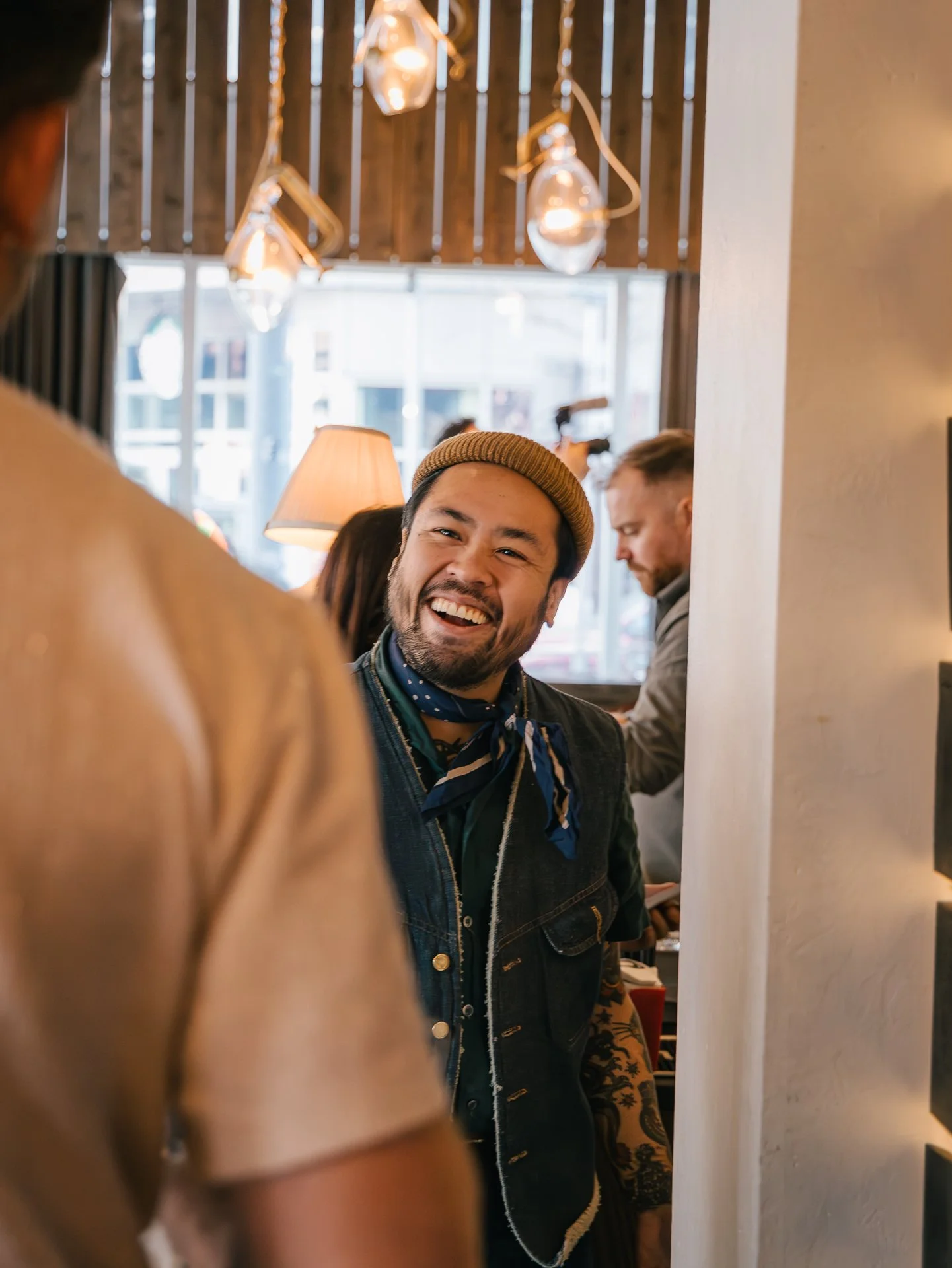 A man with a beard and mustache, wearing a tan beanie, smiling and looking to his right inside a cozy, well-lit cafe with hanging lights and large windows.