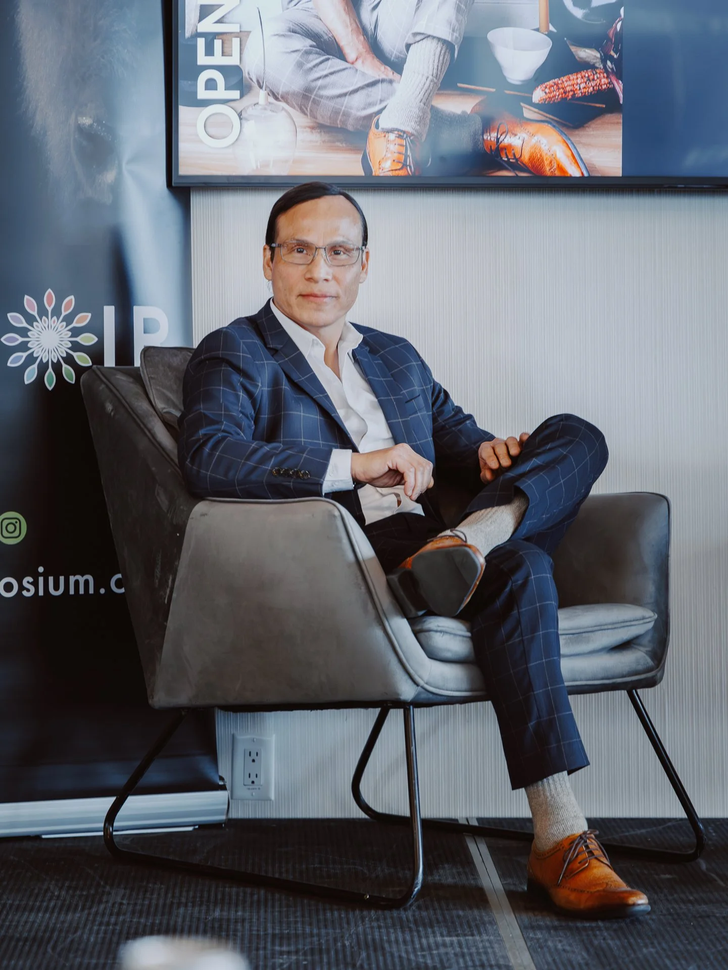 A man in a business suit sitting in a modern chair, wearing glasses, and smiling at the camera. Behind him is a large poster with the word 'OPEN' and some images of food and drink.