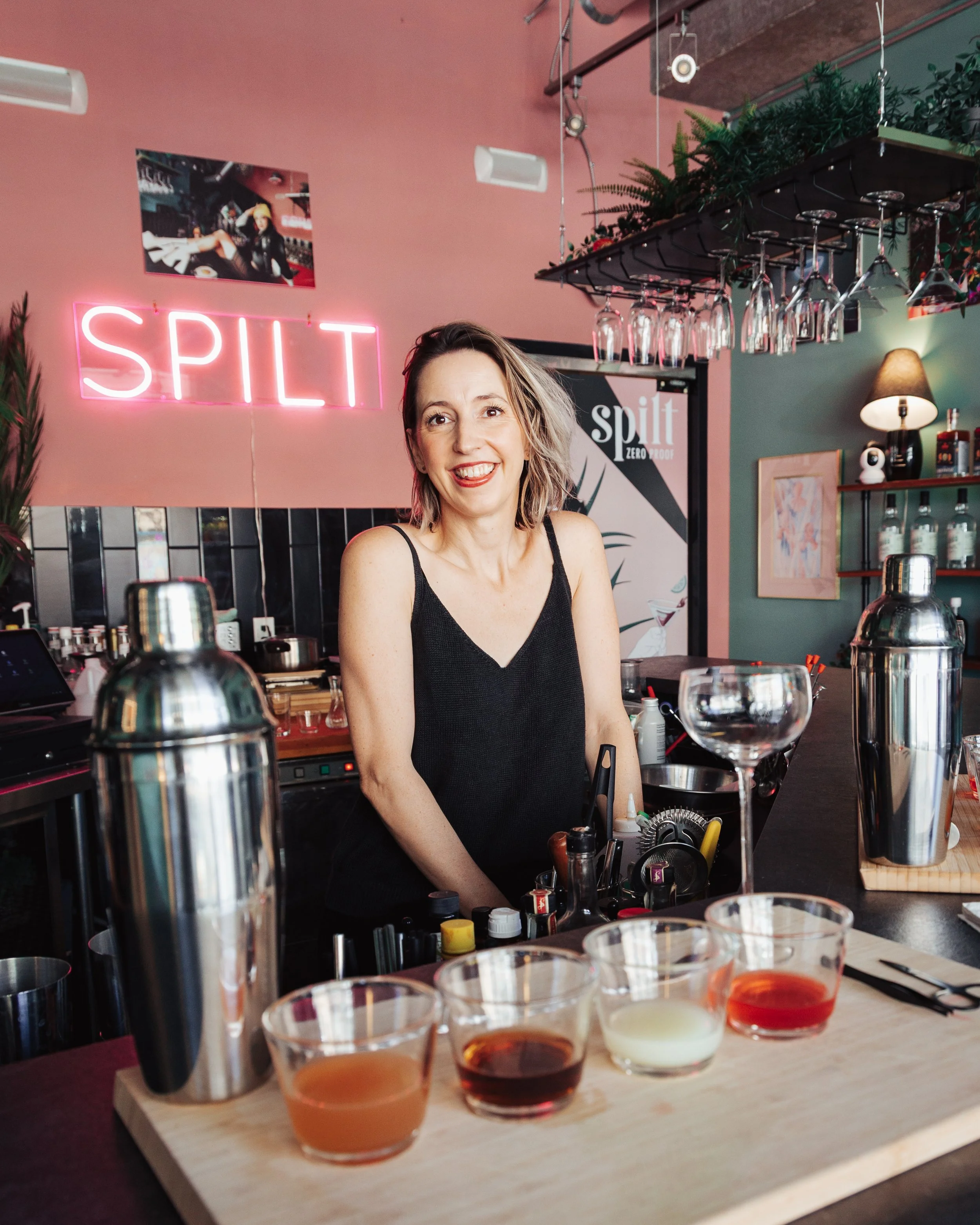 A woman with short, wavy hair and a black sleeveless top sits behind a bar counter with drinks and bar tools in front of her. The background features pink walls, a neon sign that reads 'SPLIT', hanging wine glasses, framed pictures, and eclectic deco